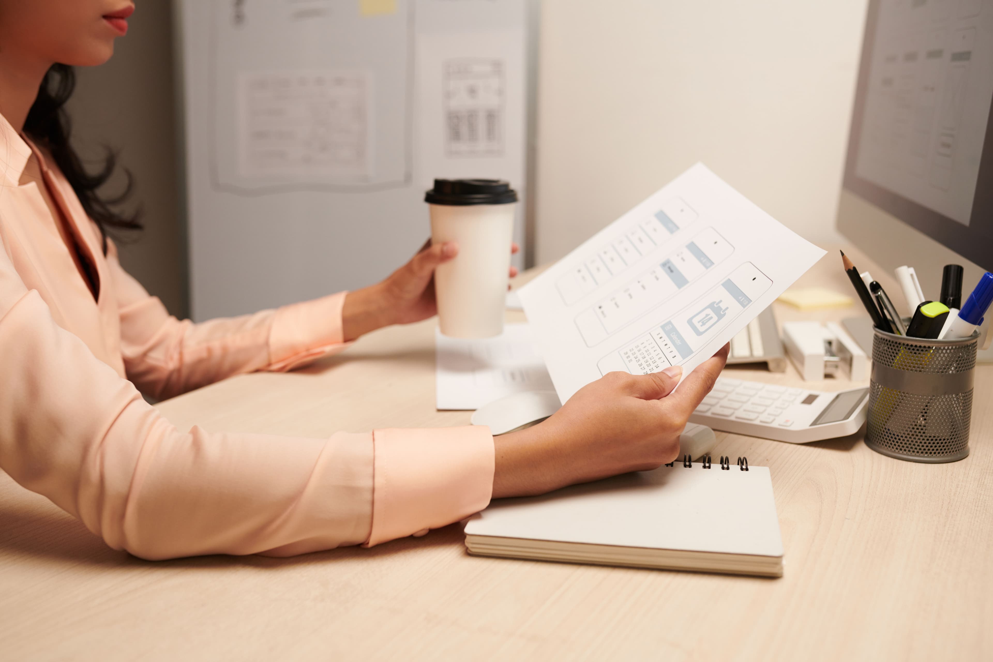 A person wearing a light blouse, holding a coffee cup and reviewing printed mockups at a desk with a computer and notepad. Suitable for "landing pages examples" for UI/UX design or professional workspace themes.