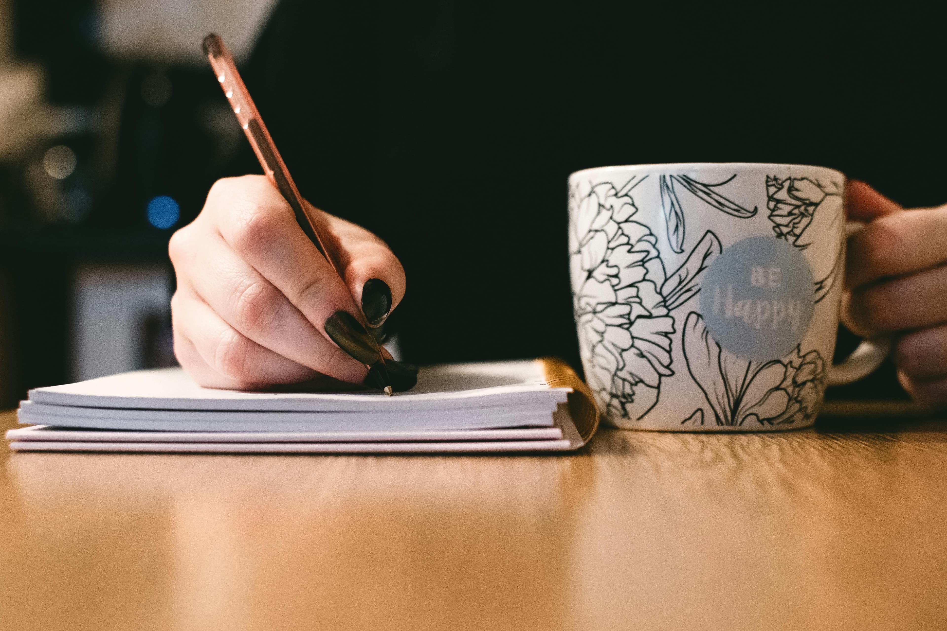 Close-up of a hand writing in a journal while holding a mug with the words 'Be Happy.' The image conveys the personal touch and mindfulness often integrated into B2B marketing automation workflows.