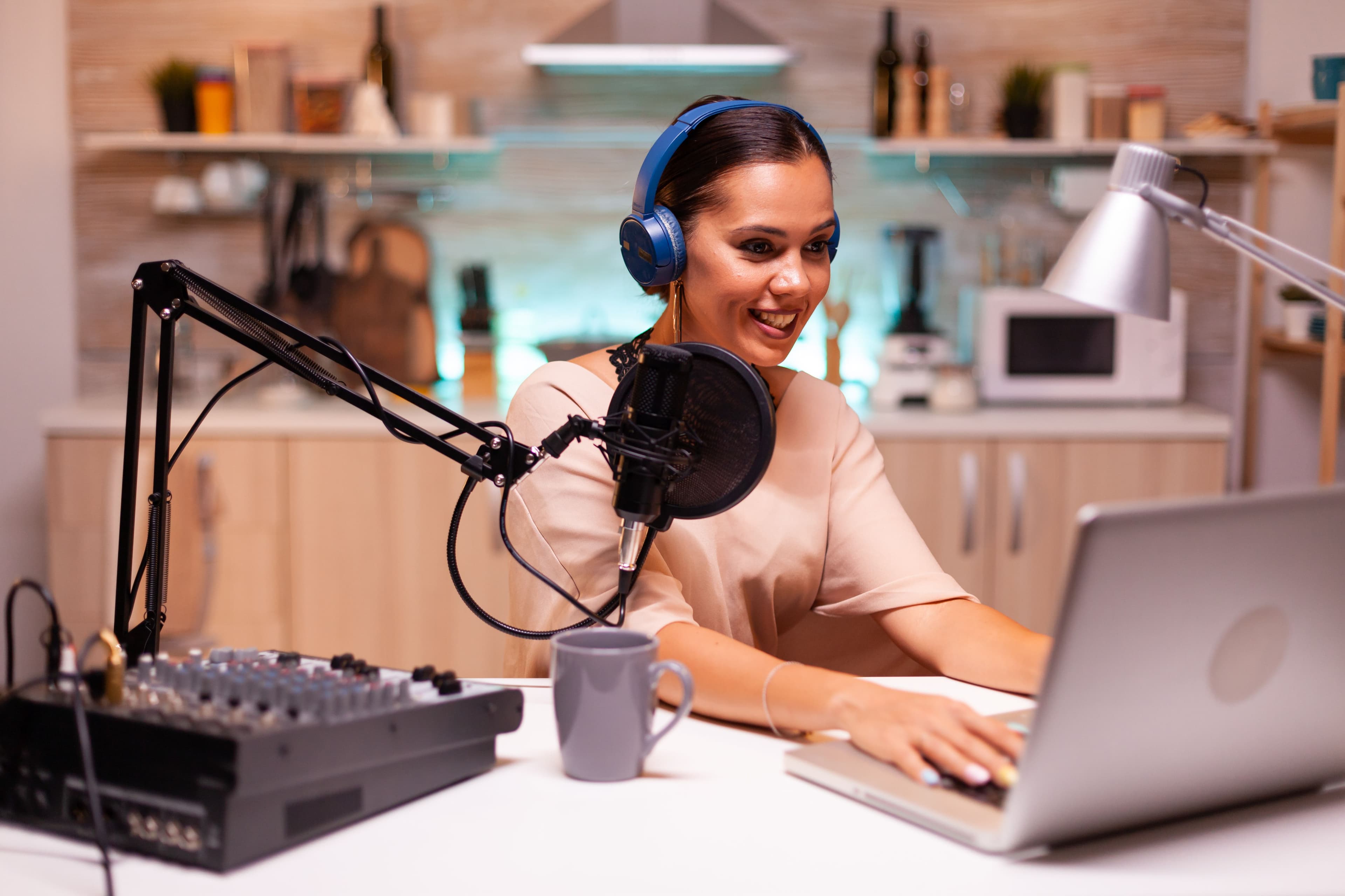 A woman wearing headphones and speaking into a microphone while looking at a laptop screen, set up with podcasting equipment in a kitchen-like background. This illustrates a "small business marketing consultant" hosting a podcast or online webinar to share marketing strategies and insights.