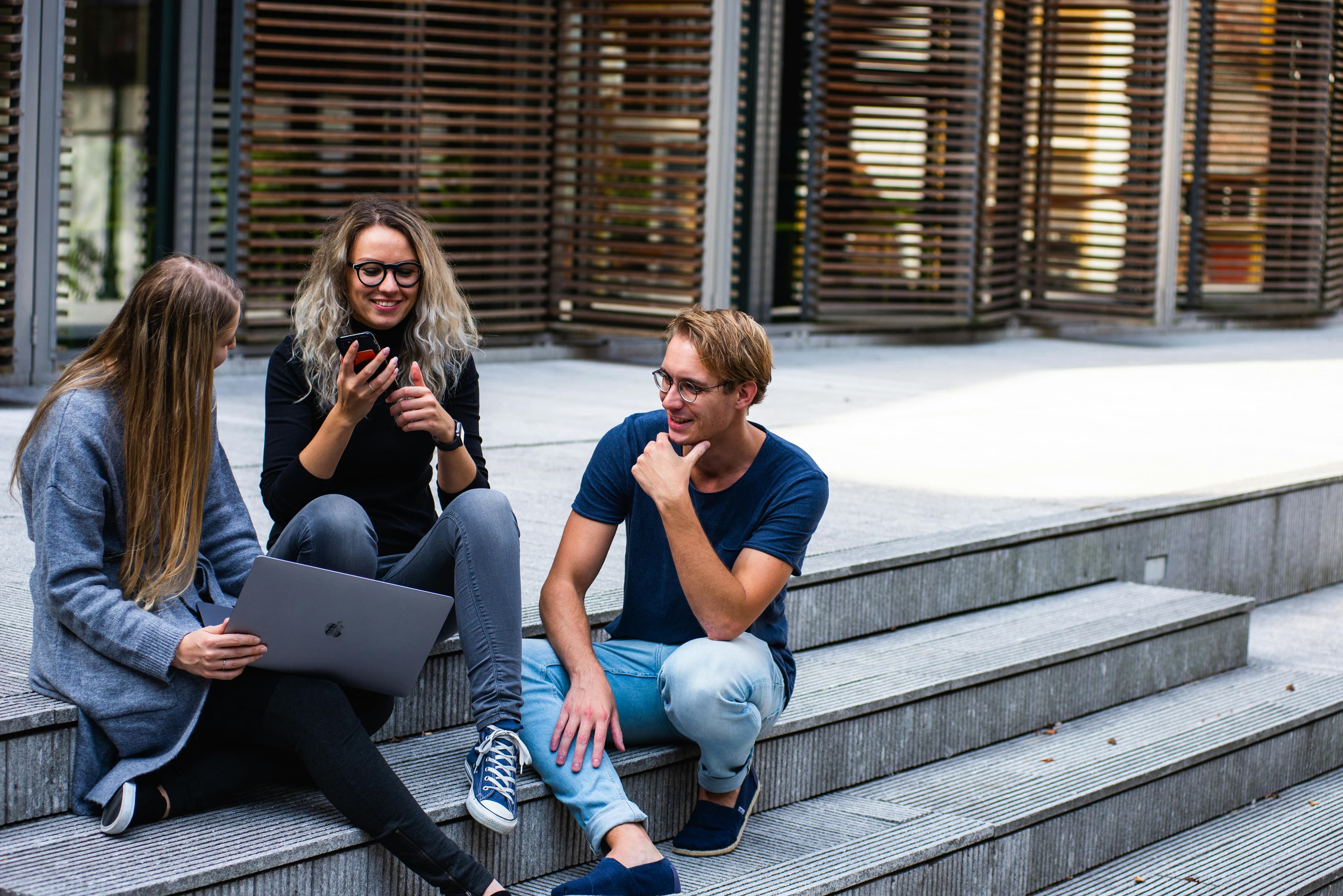 A group of three young professionals sitting on outdoor steps, laughing and collaborating while using a smartphone and laptop. This scene illustrates teamwork and creativity, emphasizing "ways to improve marketing performance" through collaboration and innovative ideas.