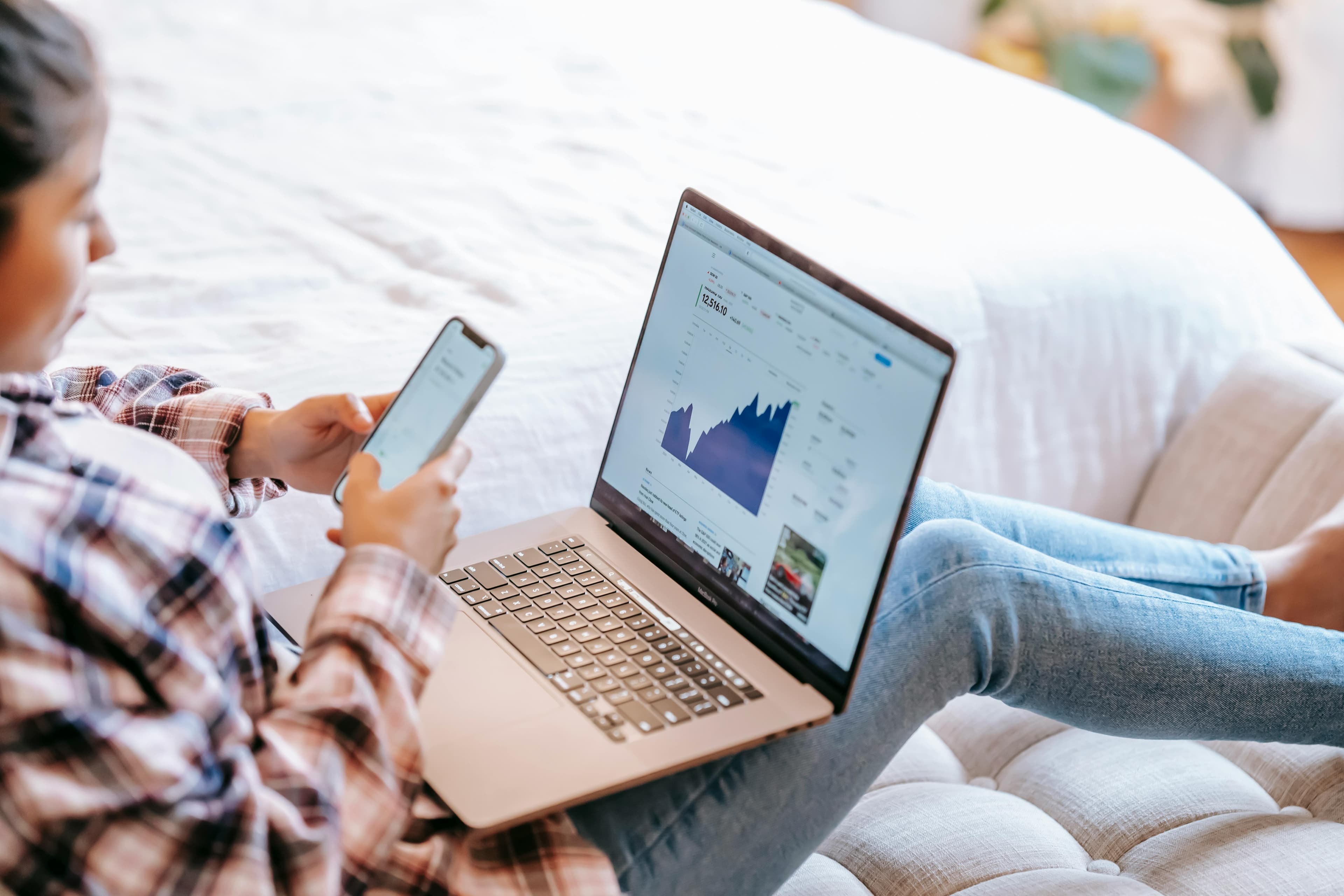 A person in a plaid shirt is sitting comfortably on a bed, using a laptop and a smartphone simultaneously. The laptop screen displays a financial graph, while the person looks at something on their phone, suggesting they are multitasking. This setting implies they might be exploring or using free AI copywriting tools to assist with their work, leveraging technology to enhance productivity and efficiency in a relaxed environment.