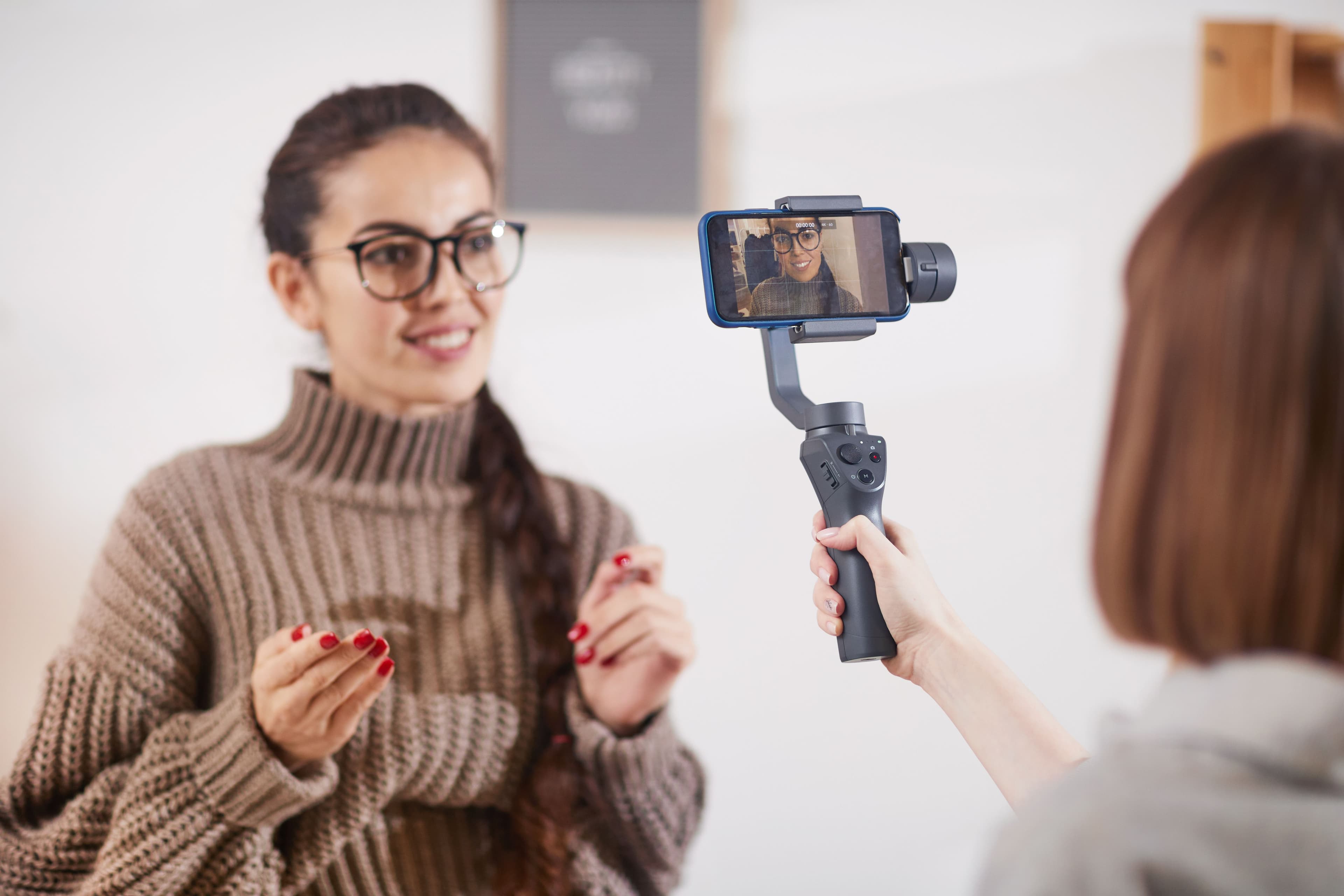 A smiling woman wearing a knitted sweater being filmed with a smartphone mounted on a stabilizer, likely creating content. Suitable for small business websites focusing on digital marketing, video production, or influencer collaborations.