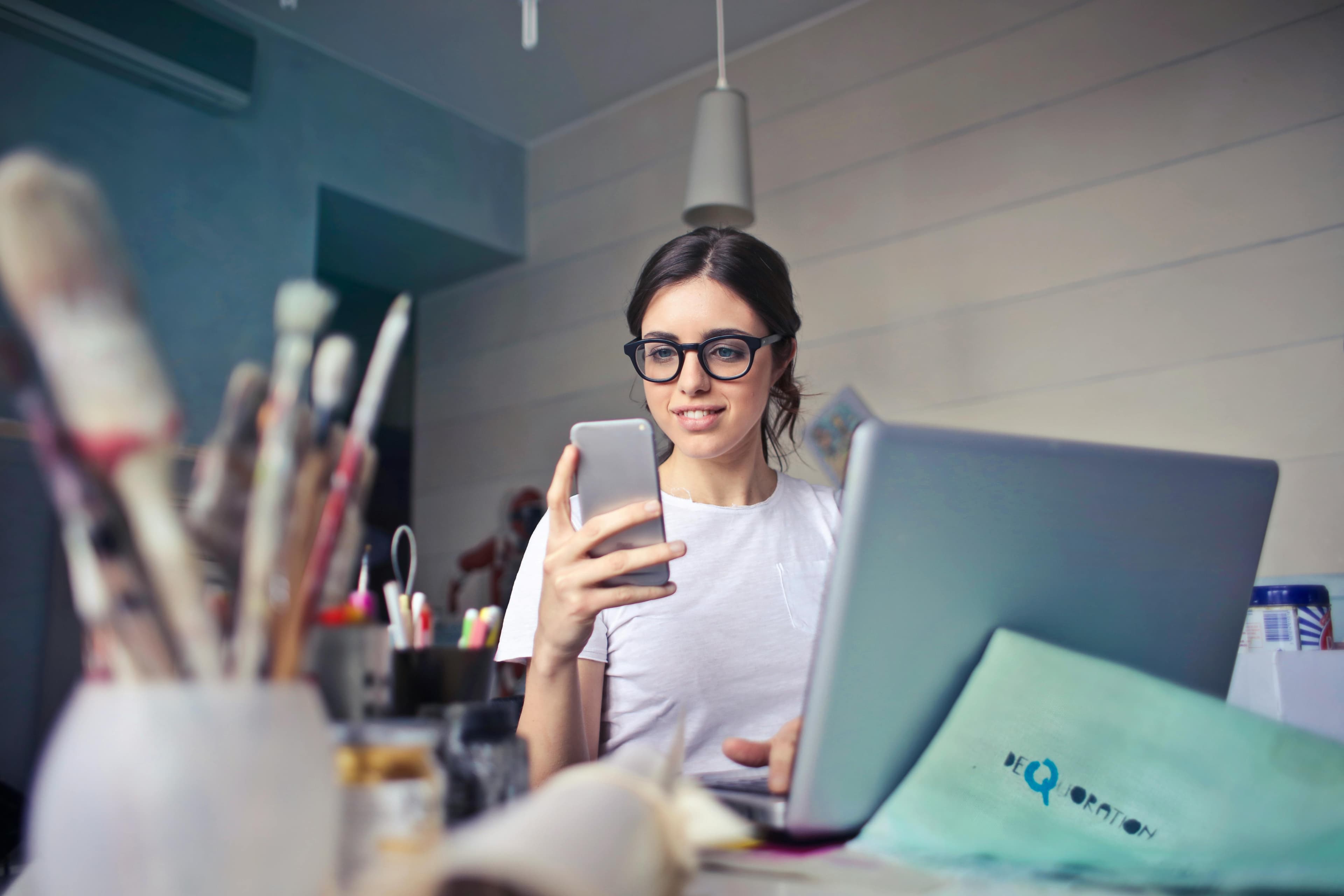 A woman wearing glasses, seated at a cluttered artist's desk, looking at her smartphone while her laptop is open beside her. This image can represent a creative professional engaging with a lead form for artistic inquiries or business.