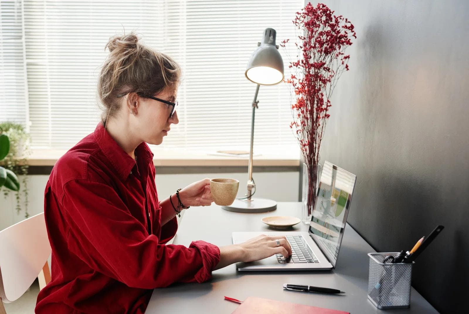 A woman in a red shirt holding a cup while preparing a page online yard sale on her laptop at a minimalist desk.