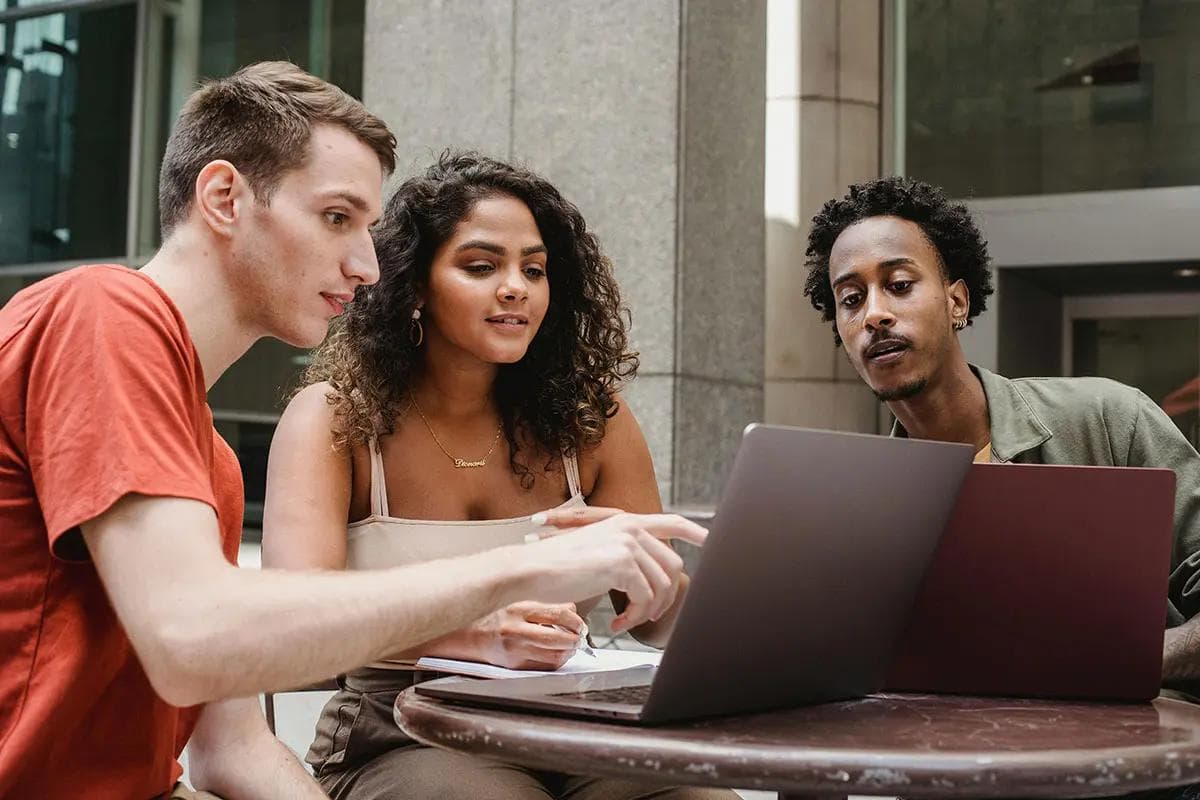 Three colleagues are engaged in a collaborative discussion around a small outdoor table. They are focused on a laptop screen, with one person pointing at the display while the others attentively observe. The setting is a professional yet casual environment, with modern architecture in the background. This scene illustrates a productive meeting, ideal for the  concept of B2B lead generation.