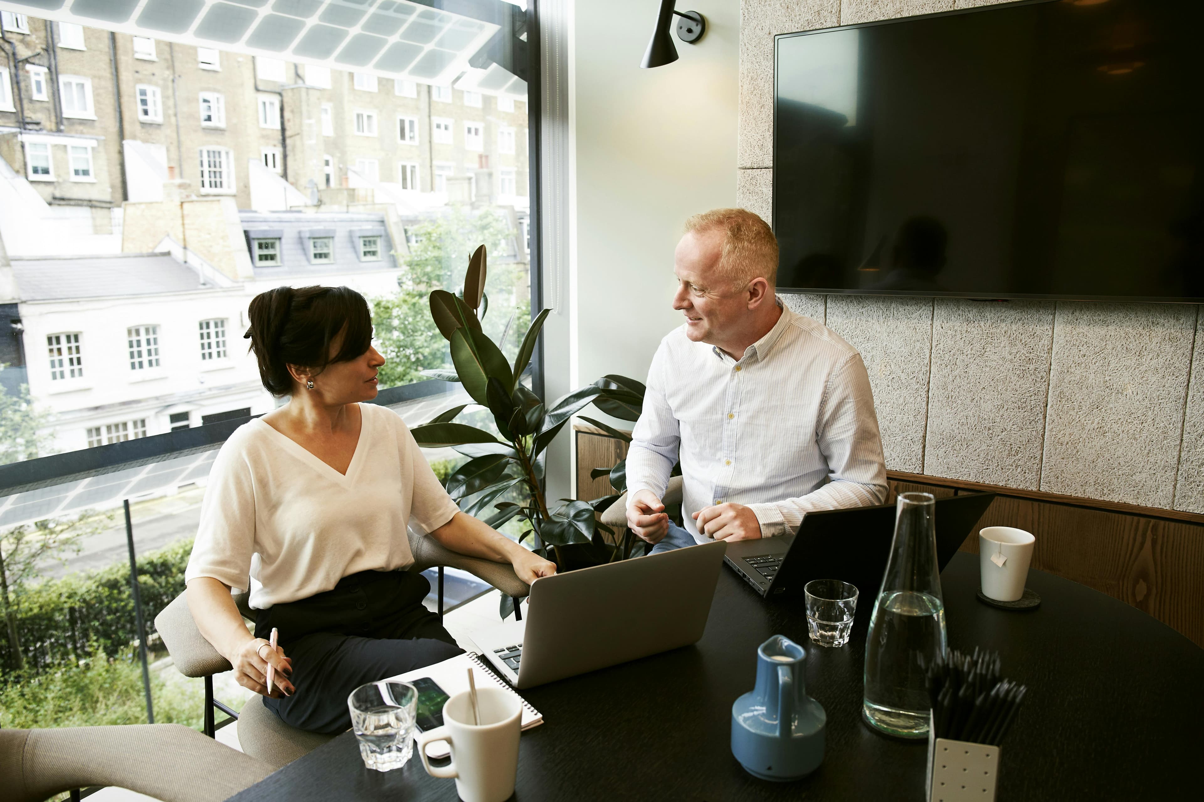  Two professionals having a discussion in a modern office setting, with a large window overlooking city buildings. The woman on the left, wearing a white blouse, is holding a pen and appears engaged in the conversation. The man on the right, dressed in a light blue shirt, is smiling and contributing to the discussion. Both have laptops open on the table, along with notebooks, glasses of water, and a coffee cup. The scene suggests a collaborative meeting at an email marketing agency, where they are likely strategizing and planning their next campaign.