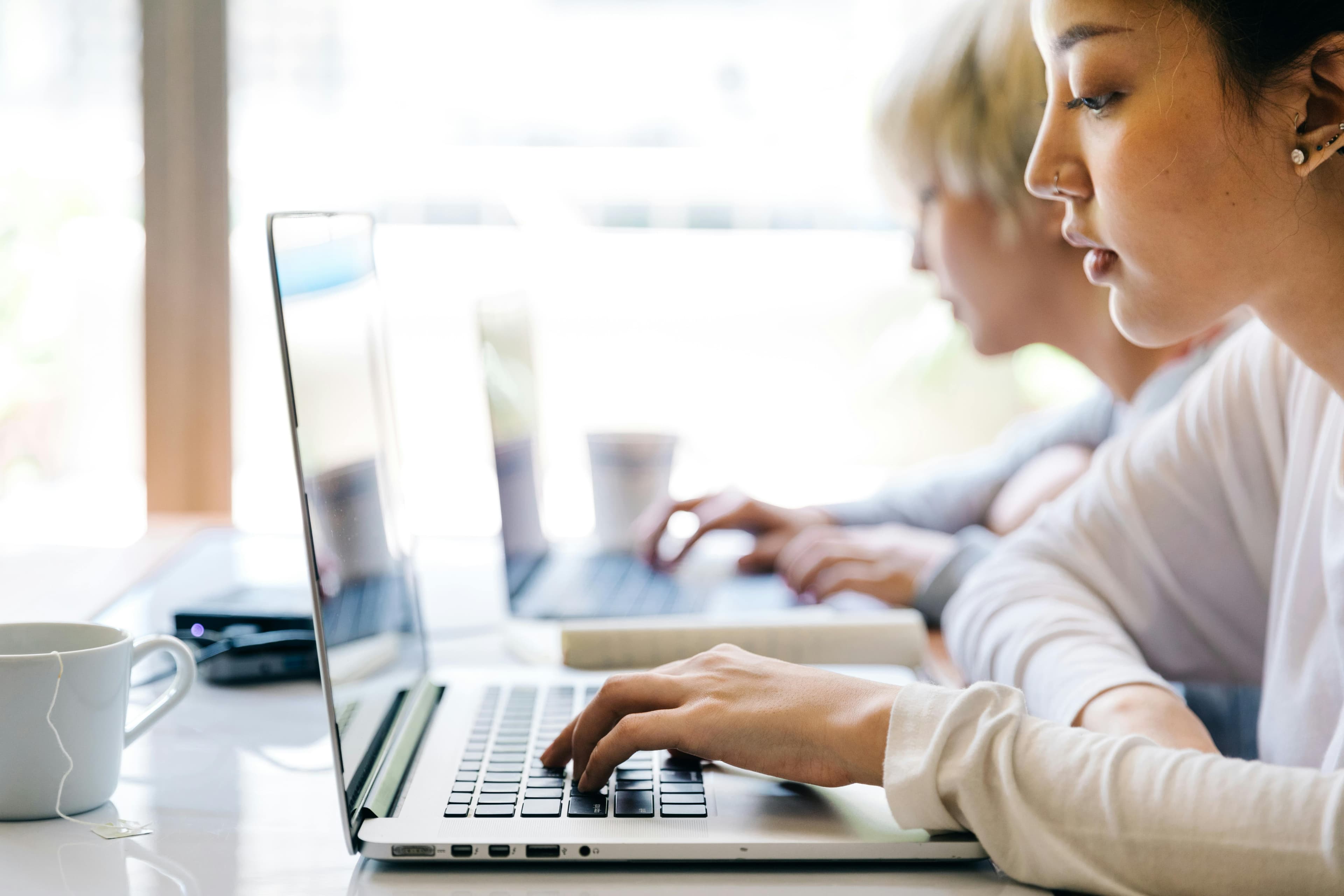 Two individuals are working intently on their laptops at a bright and airy workspace. The person in the foreground is focused on typing, with a cup of tea beside them. The collaborative environment suggests a modern office or co-working space. This scene represents the use of generative AI in marketing, where team members are likely utilizing advanced AI tools to create and optimize marketing content and strategies efficiently. The natural light and minimalist setup promote a productive and innovative atmosphere.