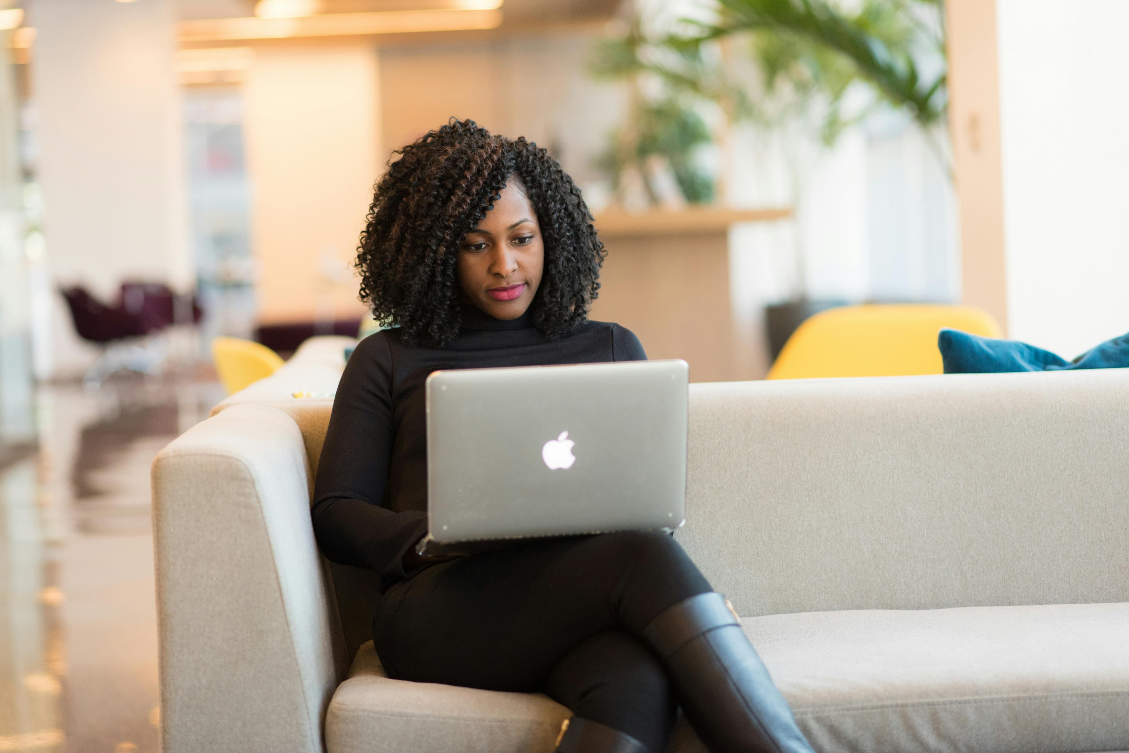 A woman sitting on a beige sofa in a modern office space, working on a silver Apple laptop. She appears focused and engaged, possibly analyzing data or strategies for eCommerce conversion optimization. The background features stylish furniture and plants, adding to the contemporary ambiance.