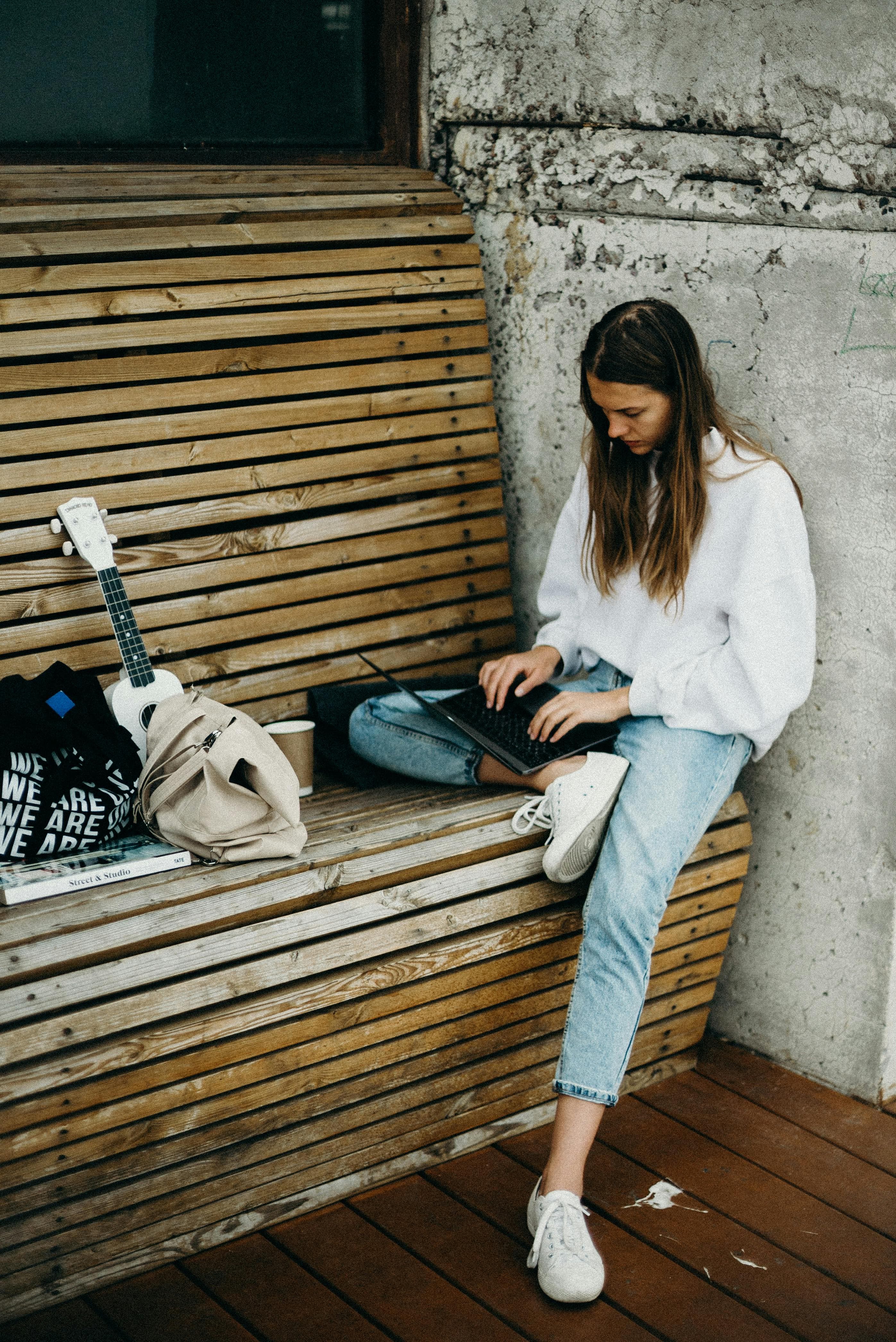  A woman sitting on a wooden bench with a ukulele, a backpack, and a laptop, engaging in focused work. This setup resonates with "action pages" that encourage creativity, freelancing, or online learning opportunities.