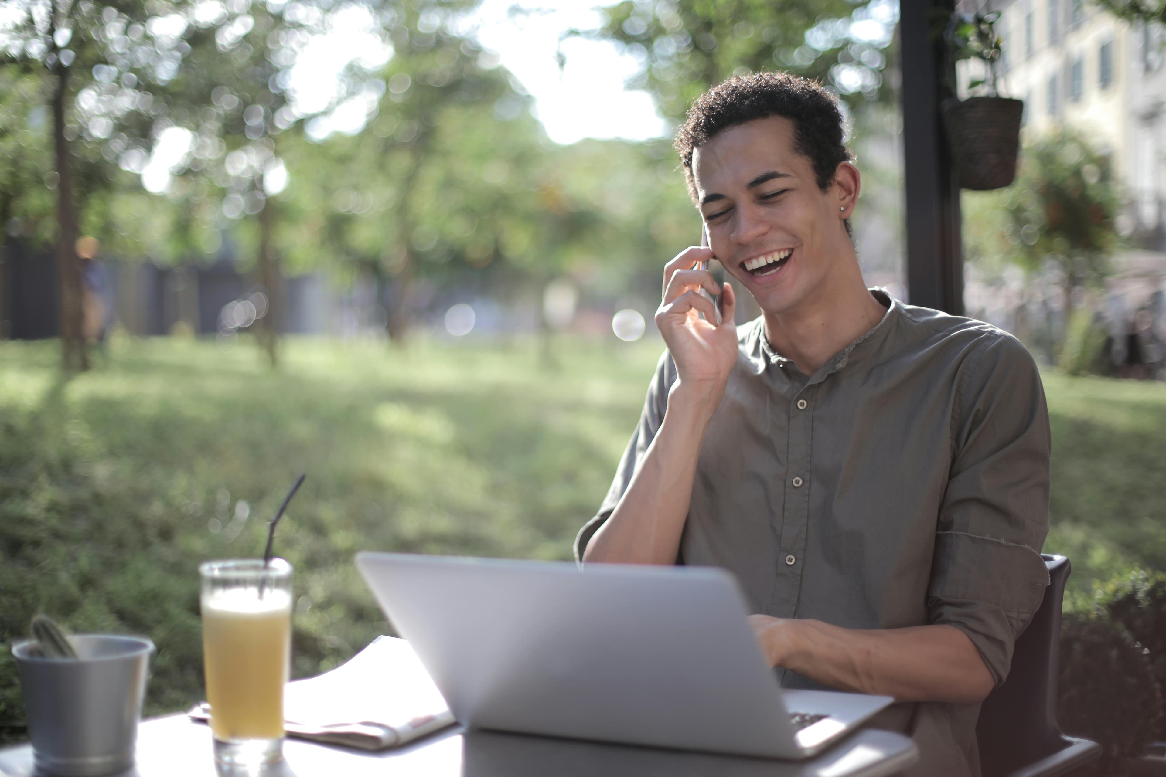 A man sitting at an outdoor cafe, smiling as he discusses strategies for optimizing product landing pages on a phone call, with a laptop and drink nearby.