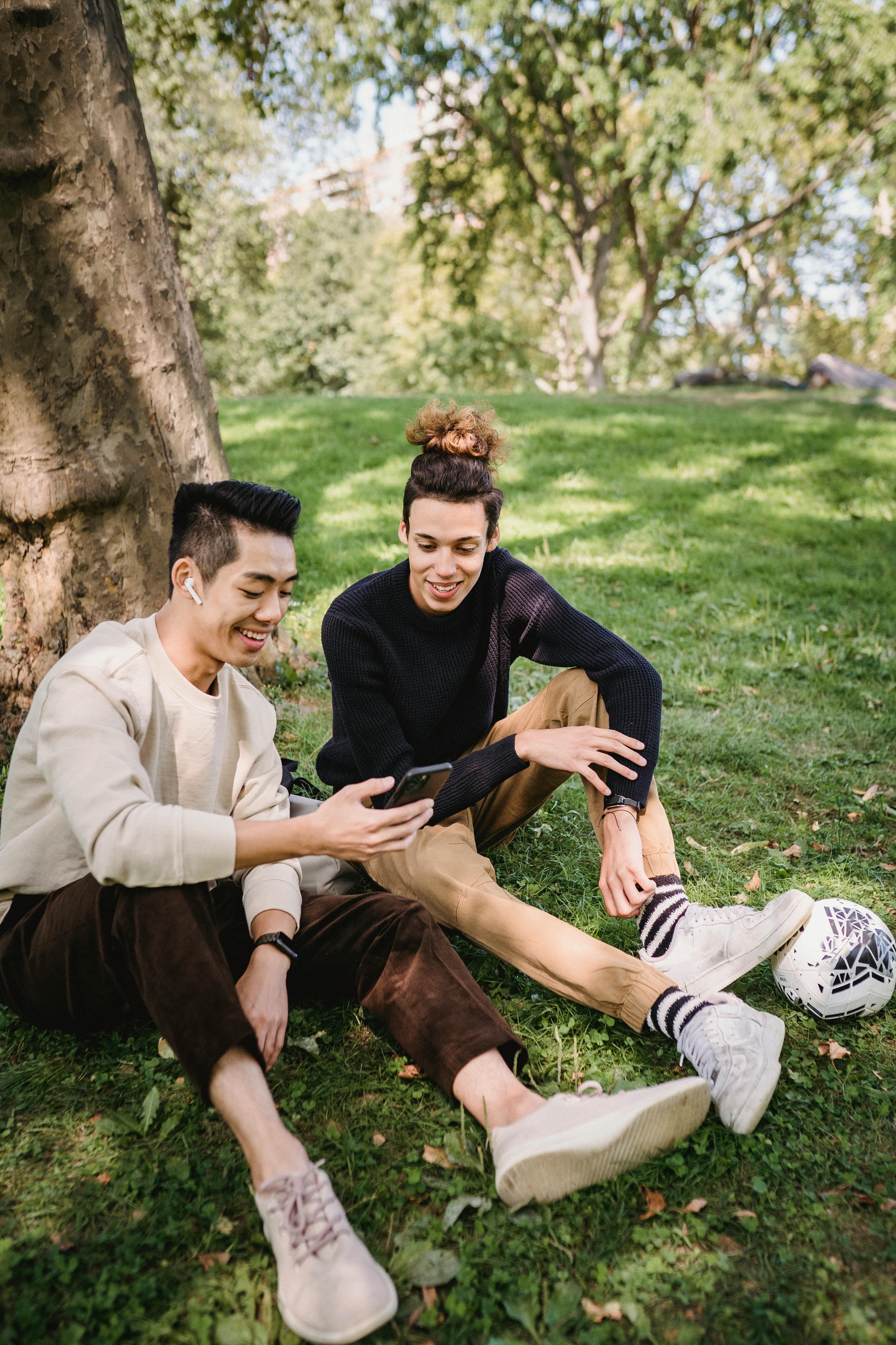 Two young men sitting on the grass in a park, looking at a phone together and discussing creative strategies on "how to design a website that converts" in a relaxed, collaborative setting.