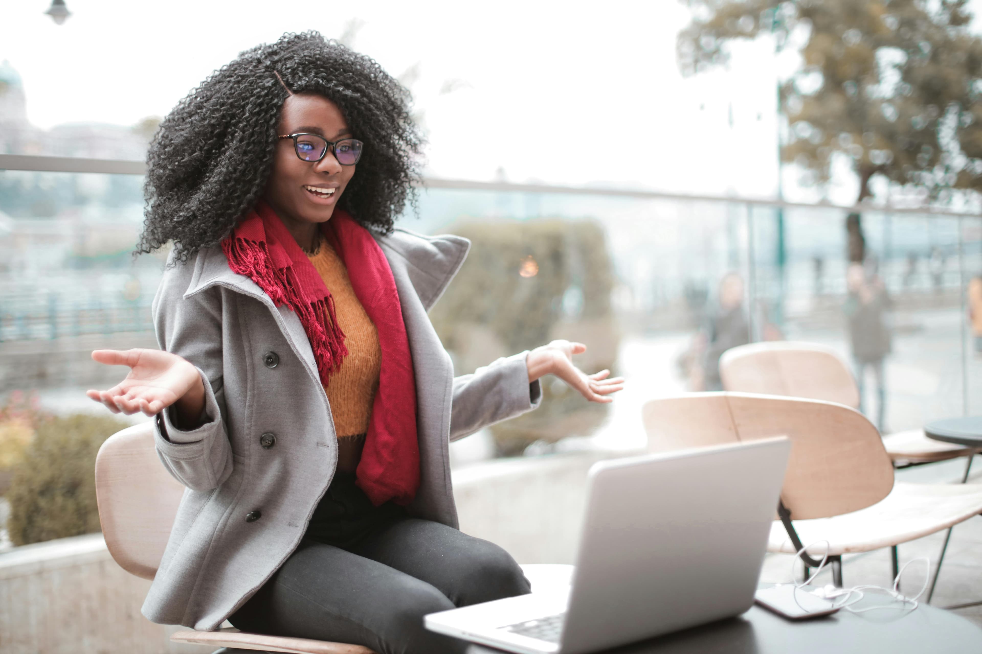 A woman with glasses and curly hair, wearing a gray coat and red scarf, engaged in a video call outdoors. She gestures expressively, possibly discussing a lead generation format with colleagues in an energetic, open setting.