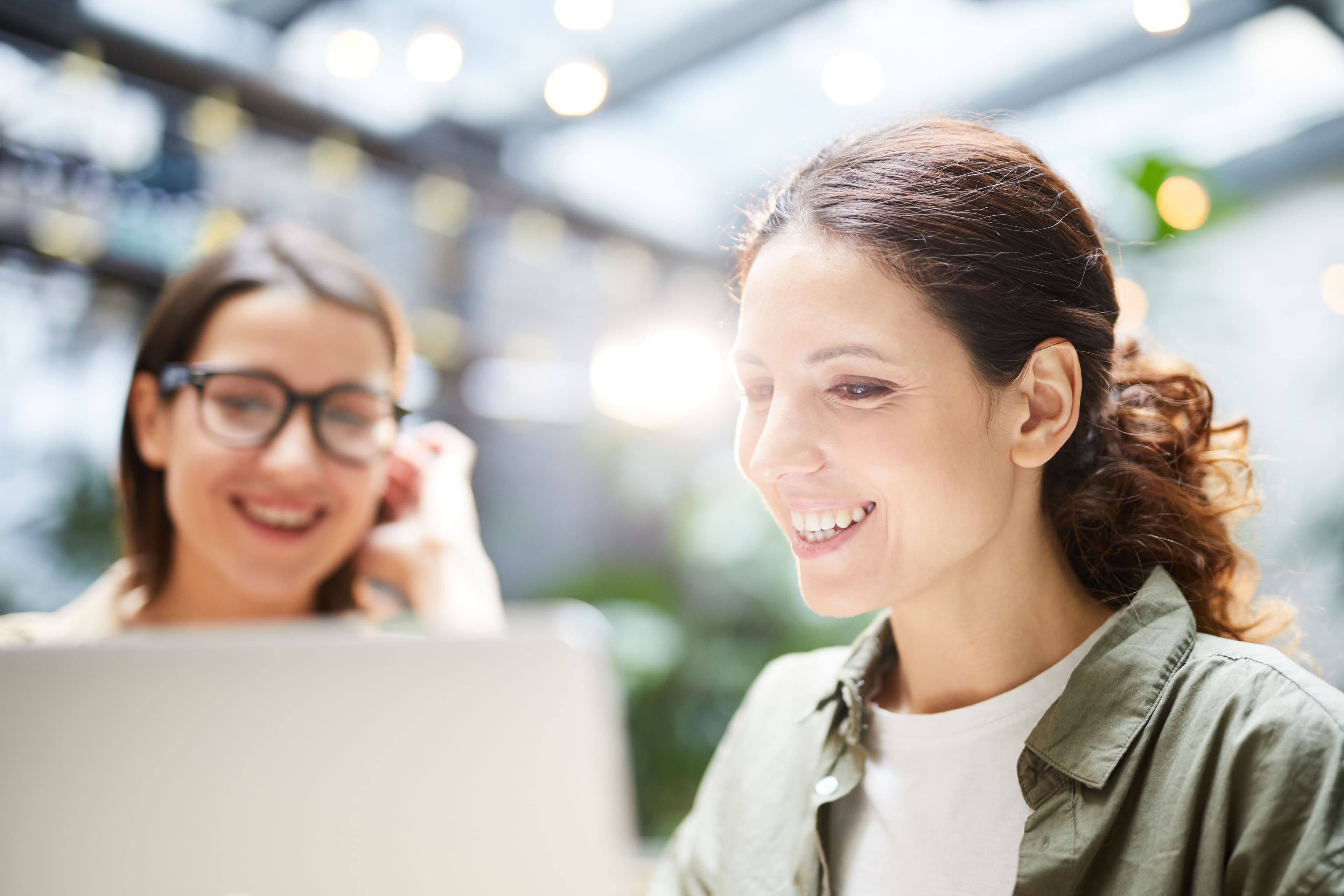  Two women collaborating in a bright, modern workspace, smiling as they review content on a laptop. This image is ideal for a landing page highlighting teamwork and preparation for a live stream product showcase.