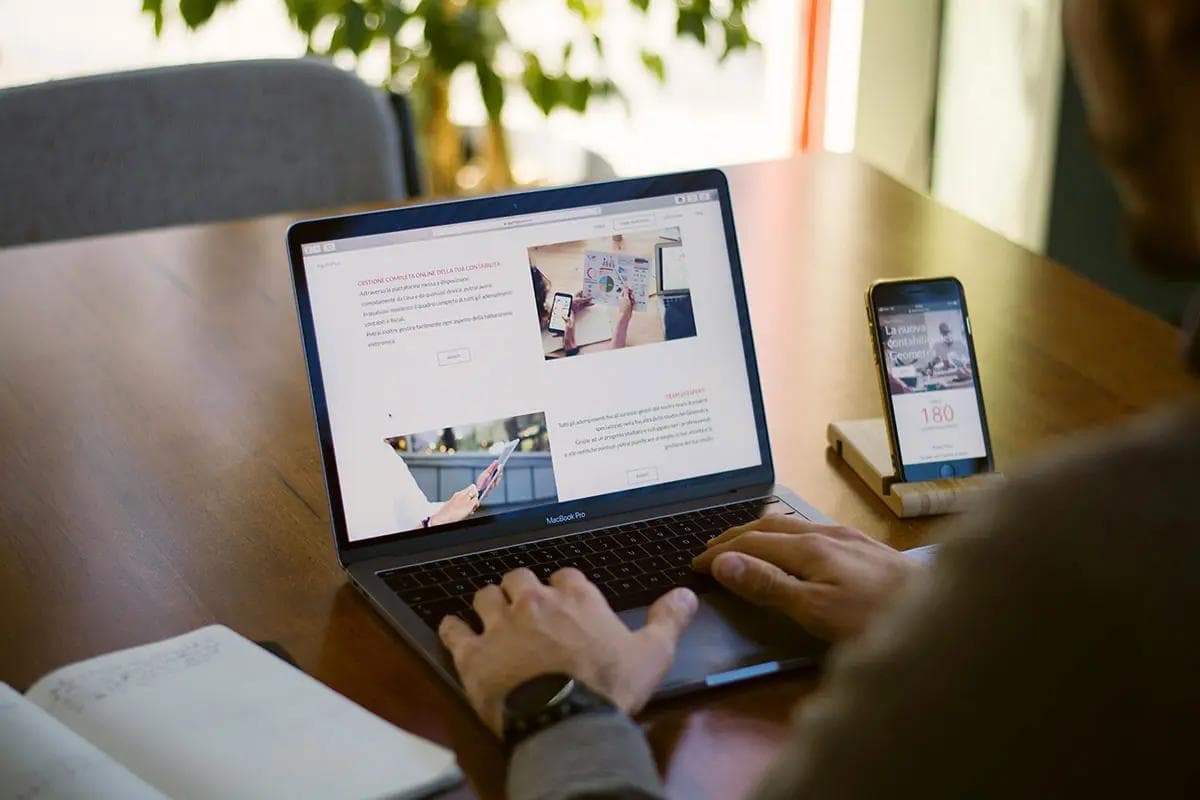 A person is working on a laptop at a wooden table, browsing a website with detailed information and images. Next to the laptop, a smartphone is propped up on a stand, displaying another webpage. A notebook with handwritten notes lies open on the table, indicating a productive work session. The background features a plant, adding a touch of greenery to the setting. This image is ideal for illustrating the concept of the "best website for small business."