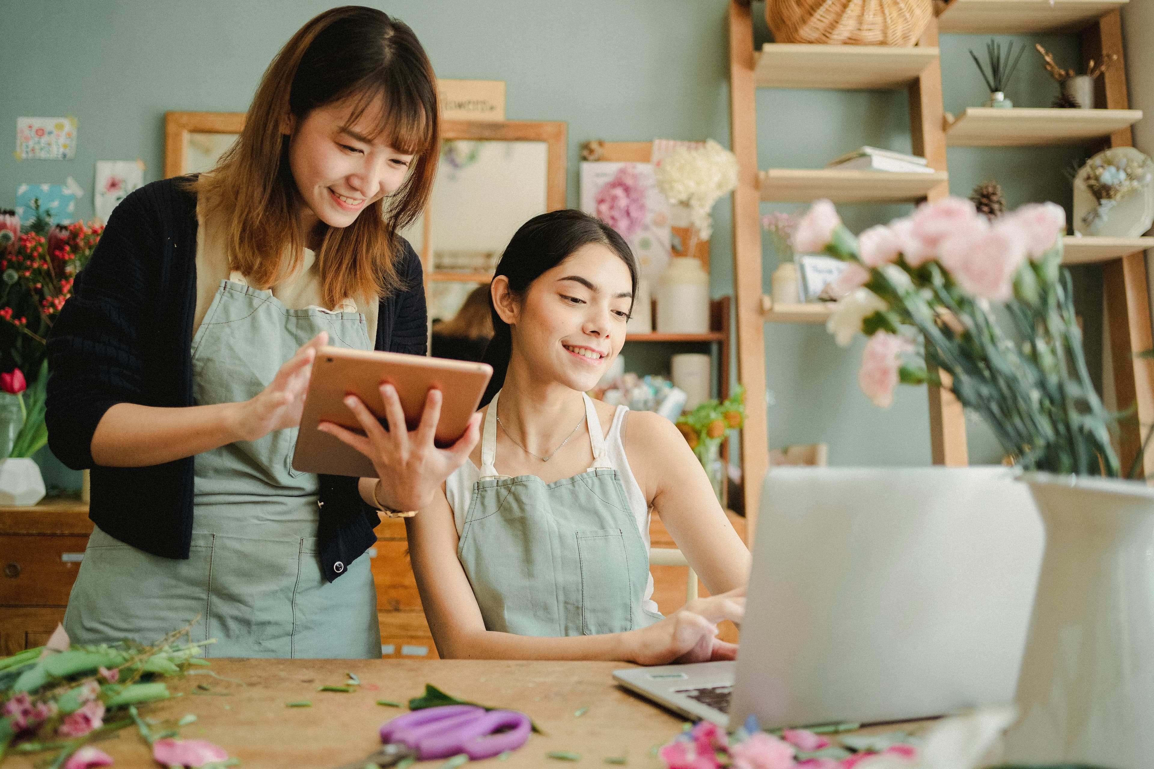 Two women in aprons collaborate on floral arrangements, smiling as they work with a tablet and laptop. This image is perfect for landing page designs showcasing teamwork, creativity, or small business success.