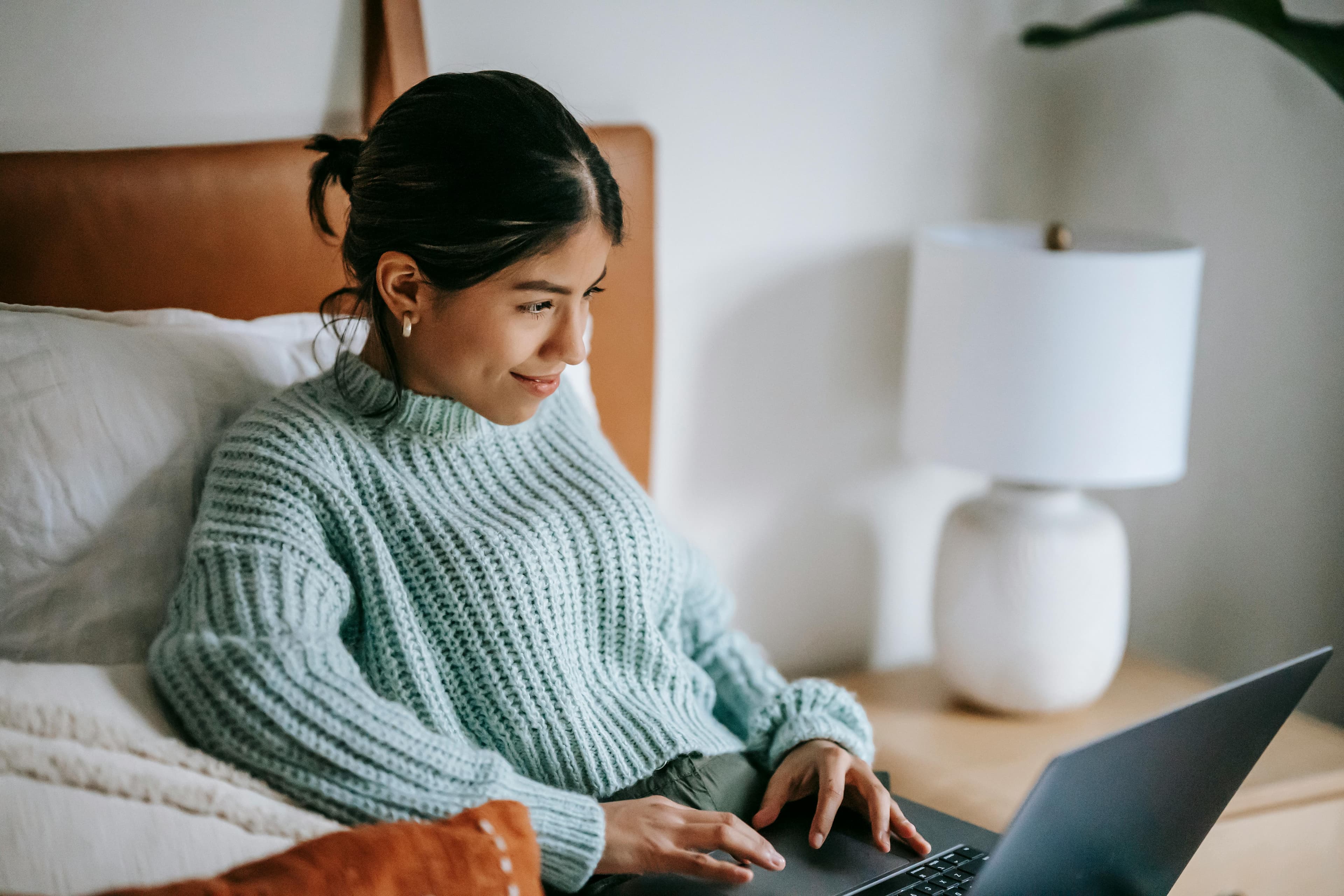 A young woman wearing a light blue sweater sits comfortably on her bed, working on a laptop. She has a content smile on her face, indicating a productive and enjoyable work session. Beside her is a nightstand with a white table lamp, contributing to a cozy and well-lit environment. This scene suggests she might be researching or analyzing trends in the Florida real estate market, taking advantage of a comfortable and serene workspace to focus on her tasks.