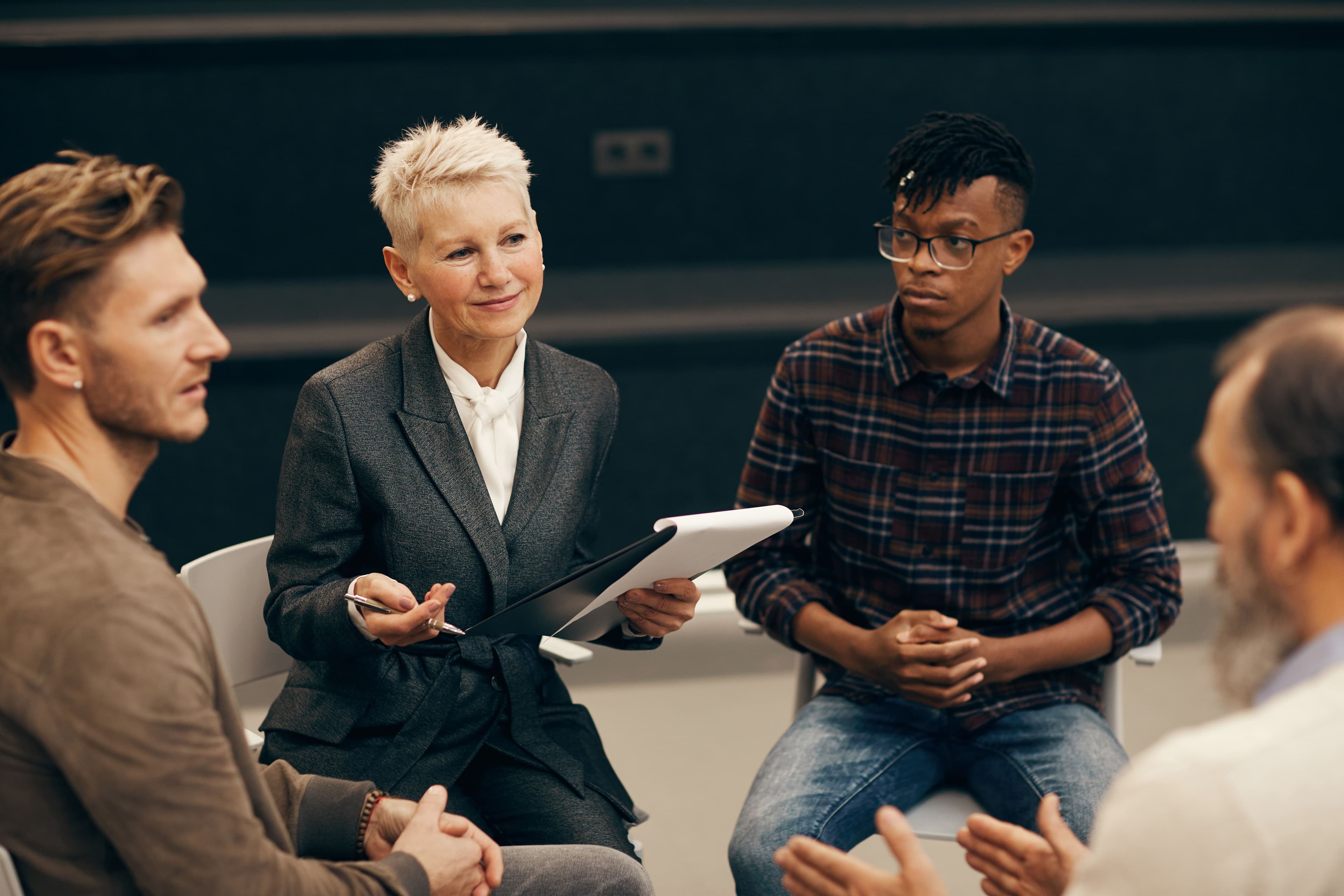 A mature woman leading a discussion in a small group setting, holding a clipboard and engaging with diverse participants. This image fits well for a landing page presenting pricing options for workshops or professional consulting services.