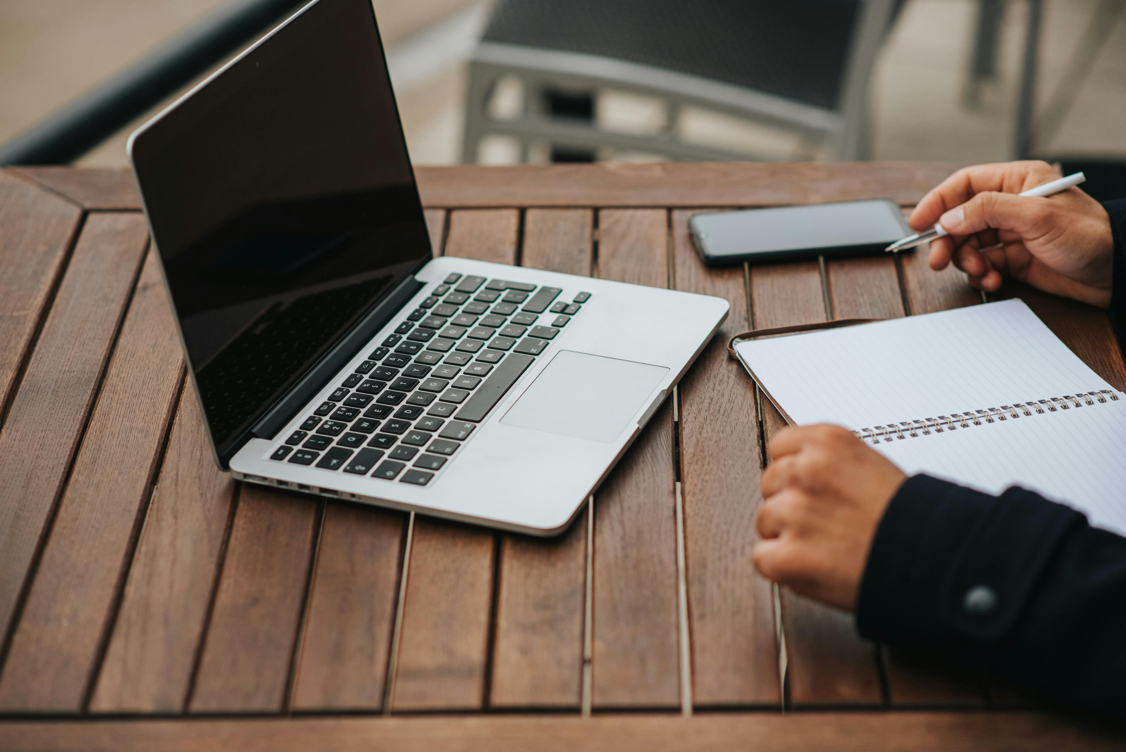 a person working on a laptop at an outdoor wooden table, with a phone and a notepad nearby. This serene setting could be ideal for researching or shopping for specialized products, such as a "lawn mower track conversion kit."