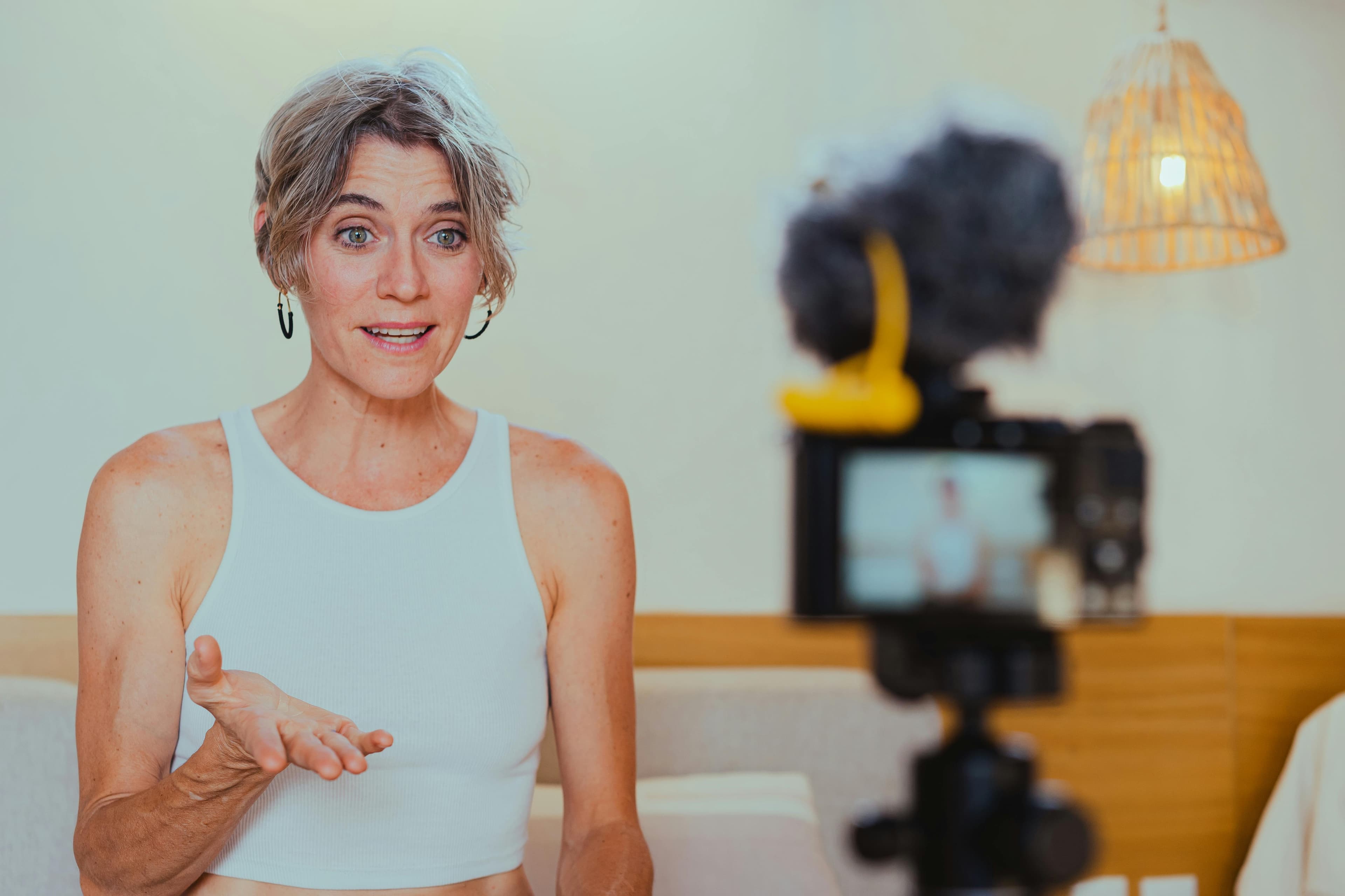 A woman wearing a white tank top is filming a video using a professional camera with a microphone setup, appearing enthusiastic and engaging, perfect for an affiliate landing page about content creation and video marketing.