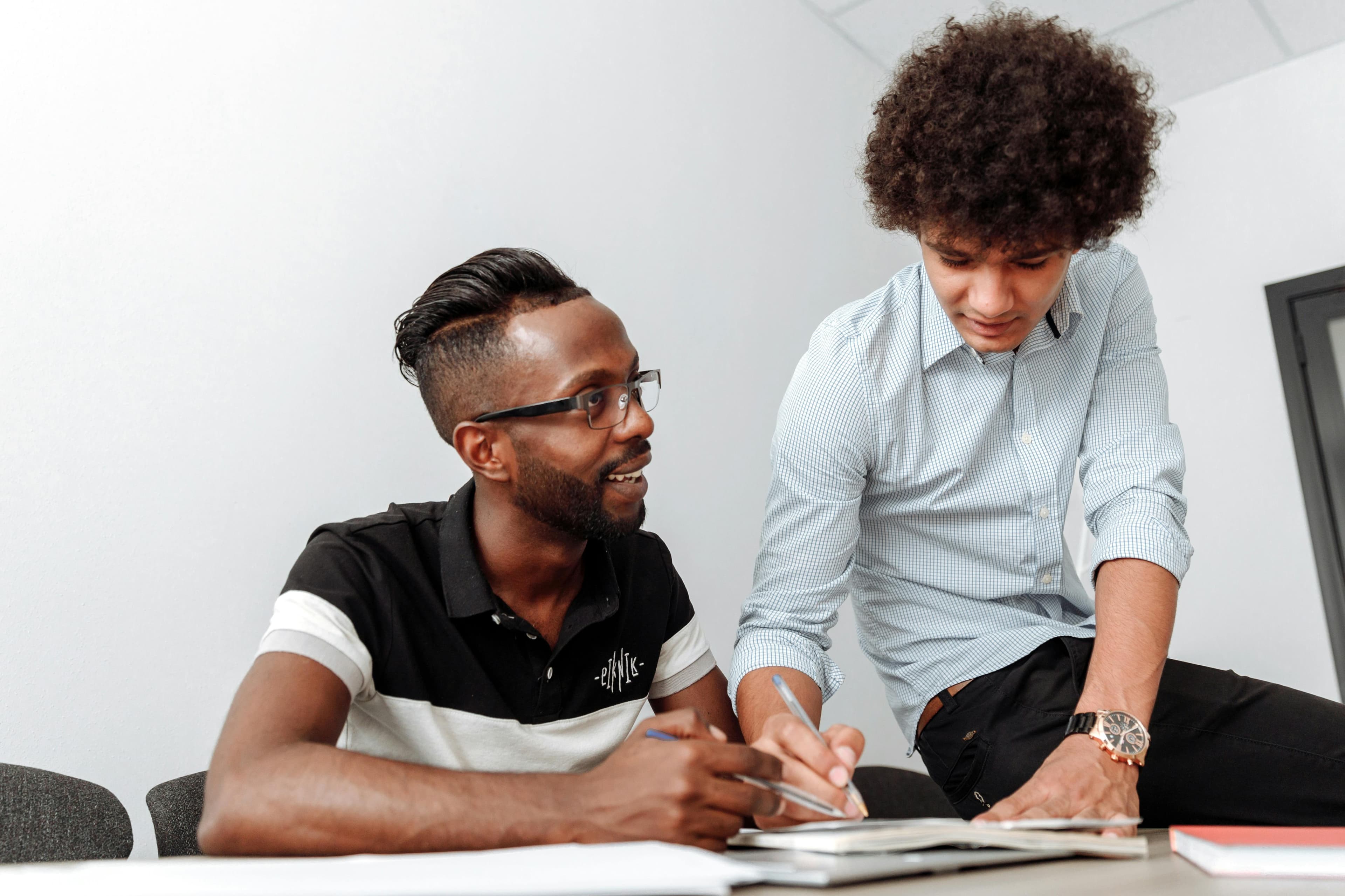 Two colleagues collaborate, one standing over the other while discussing notes. Great for illustrating teamwork and collaborative decision-making, such as setting up experiments or analyzing results using an AB testing tool.