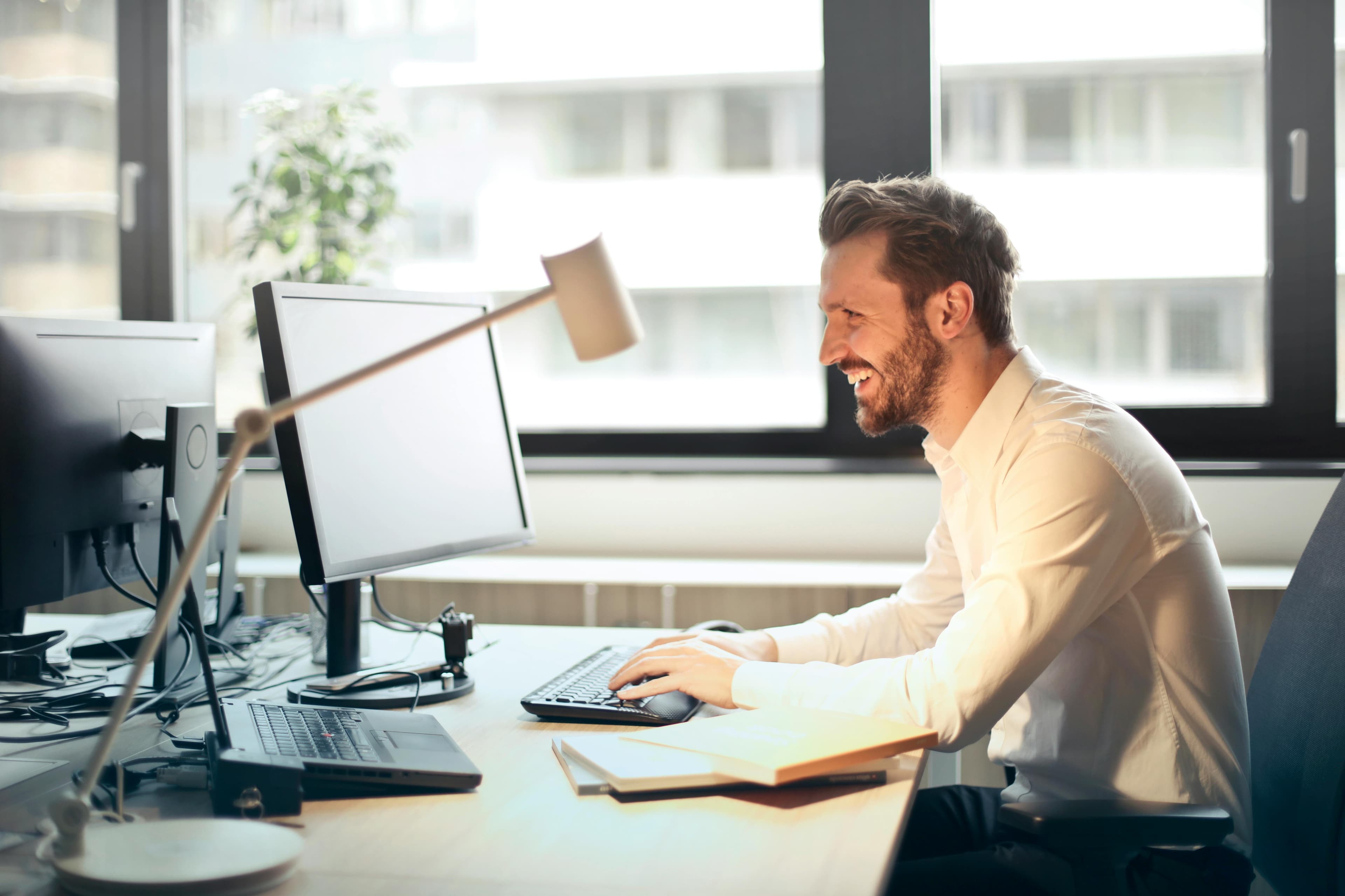 A man smiling while working at a computer desk in a modern office, perfect for a UX landing page promoting user-friendly interfaces and lead form templates for professionals.