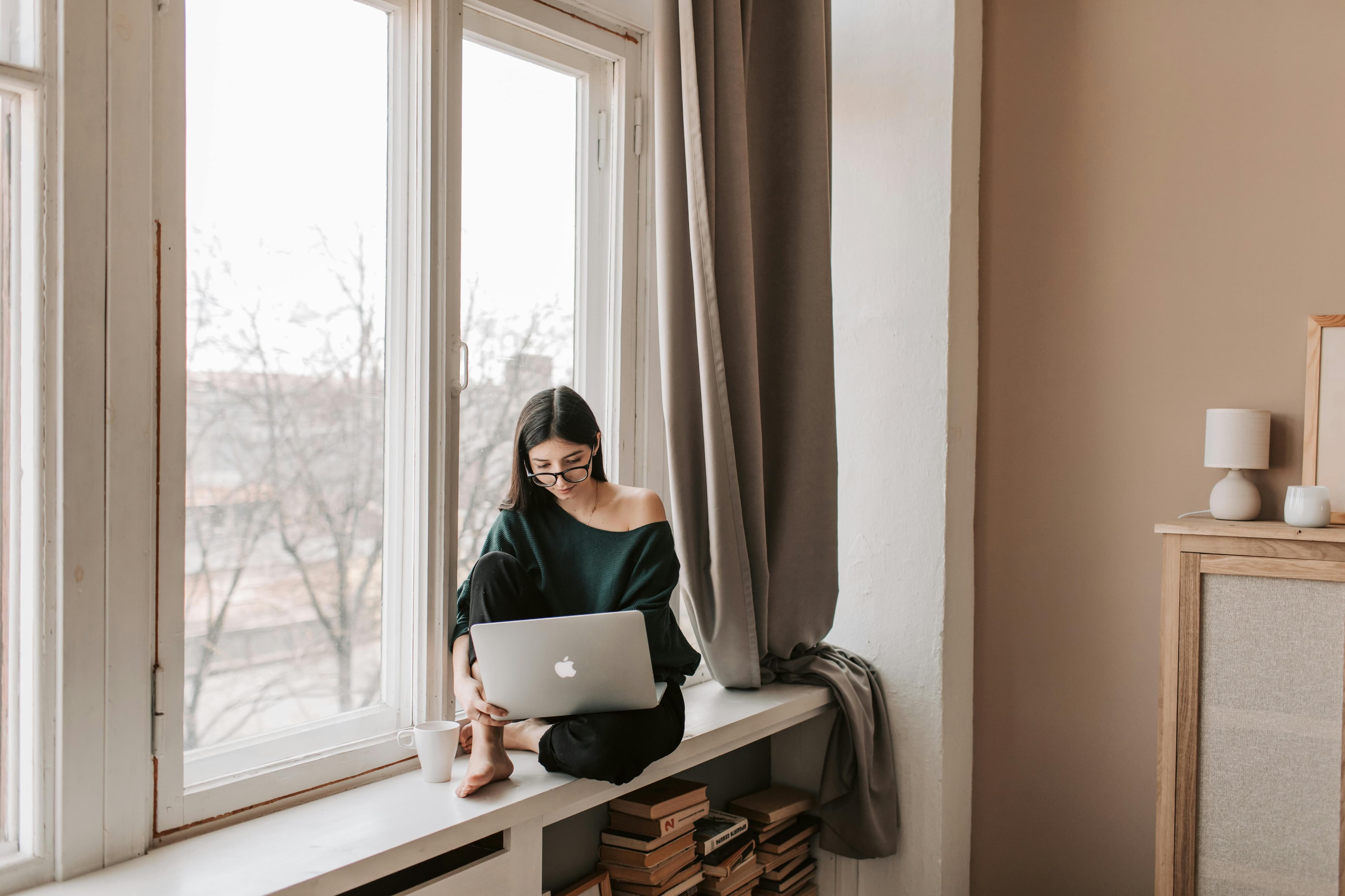 Woman sitting by a large window working on a laptop with a cup of coffee beside her, representing a copywriter vacancy.