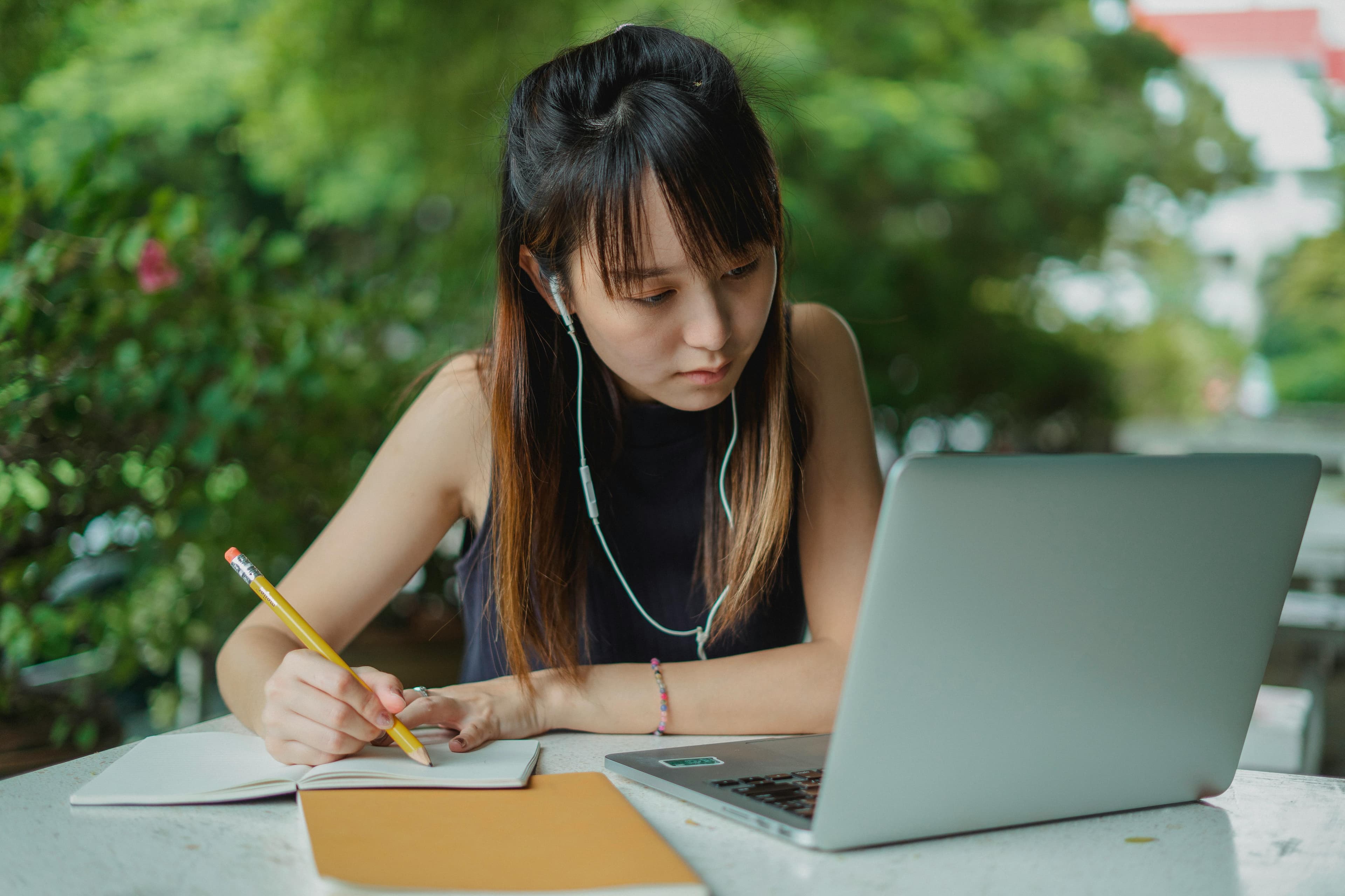 A young woman working on her laptop while taking notes in a notebook, set in a peaceful outdoor environment. This image is perfect for illustrating the "leader VS manager" concept, emphasizing a leader's ability to work efficiently in various settings.