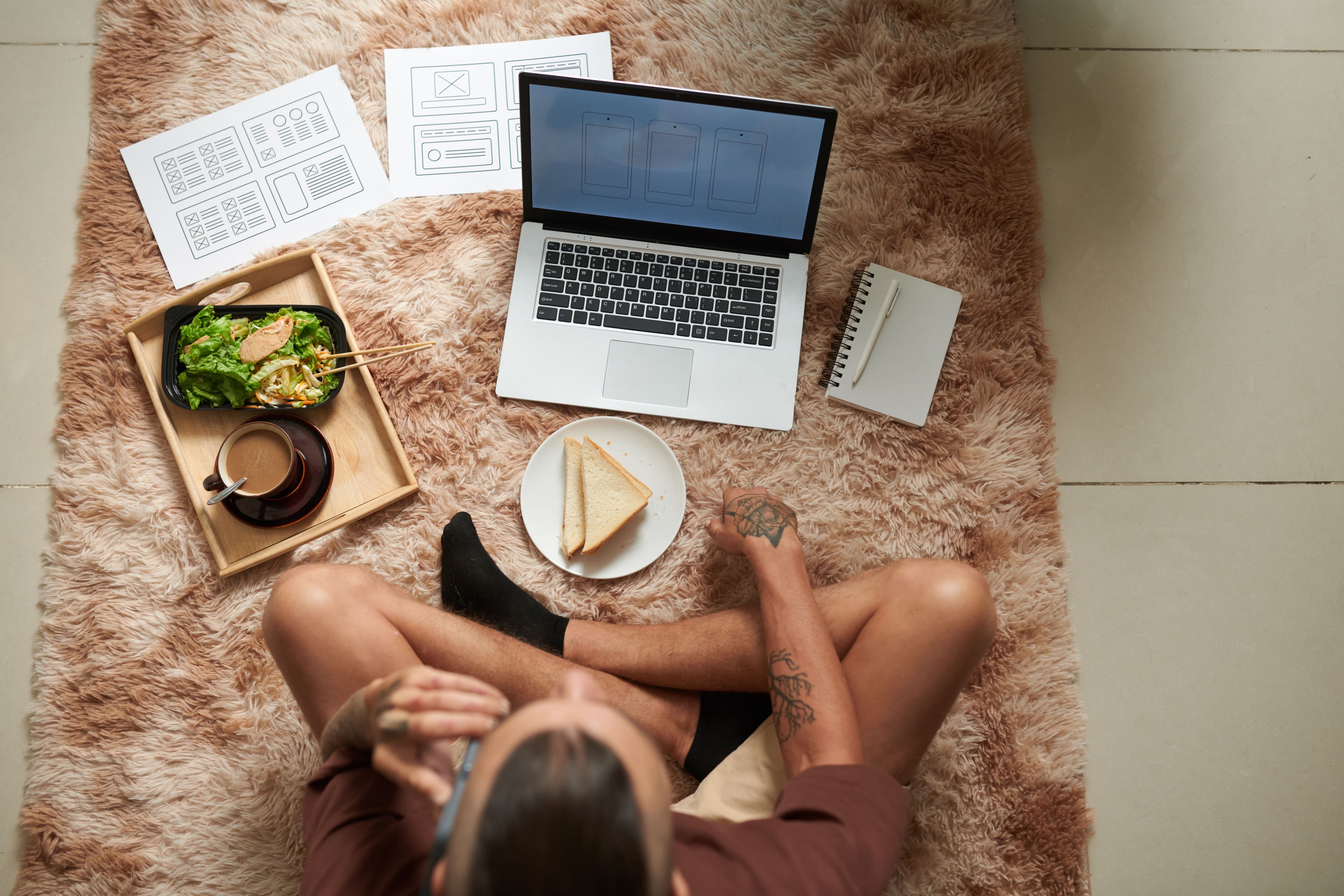 A young man working remotely from home with a laptop, surrounded by design sketches, food, and coffee, representing the flexible work environment of a search engine marketing consultant.