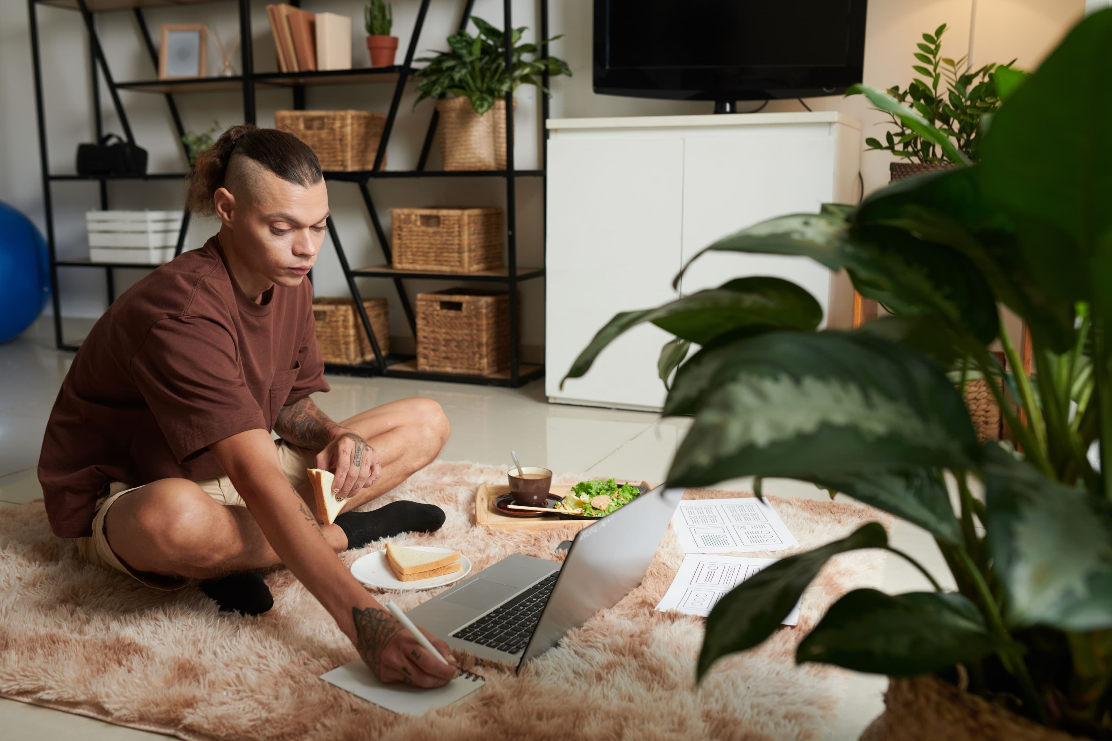 A freelance designer sitting on a rug, working on his laptop while writing down notes, possibly tracking calculator conversion metrics to measure the success of his design projects.