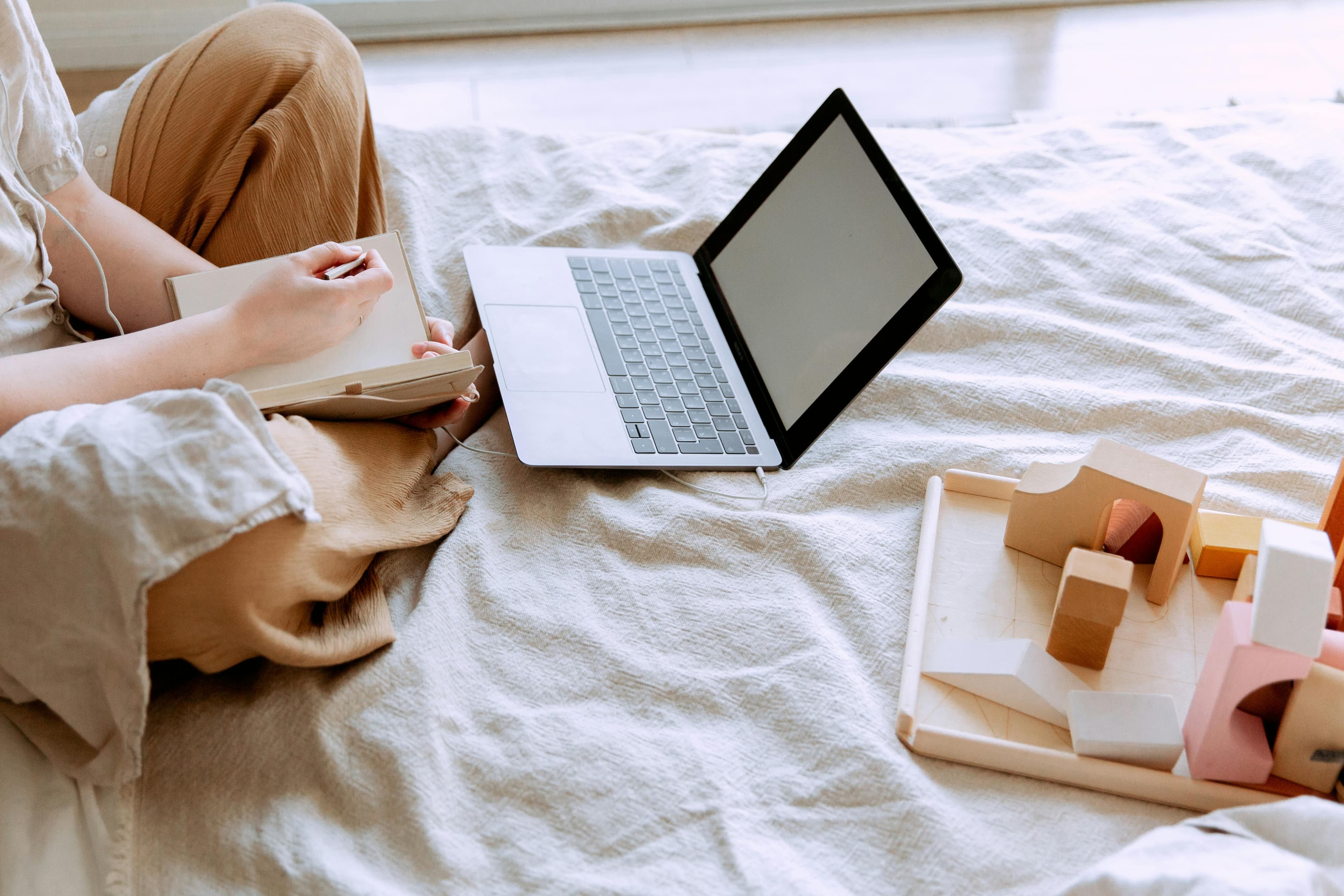 A person sitting cross-legged on a bed, working with a laptop and writing in a notebook. The scene includes a wooden toy set placed nearby, indicating a comfortable and creative workspace. This setting likely represents someone working remotely, possibly for Epic 5 Marketing and Promotions Agency, as they plan and strategize promotional activities. The natural light and cozy environment contribute to a productive and relaxed atmosphere.