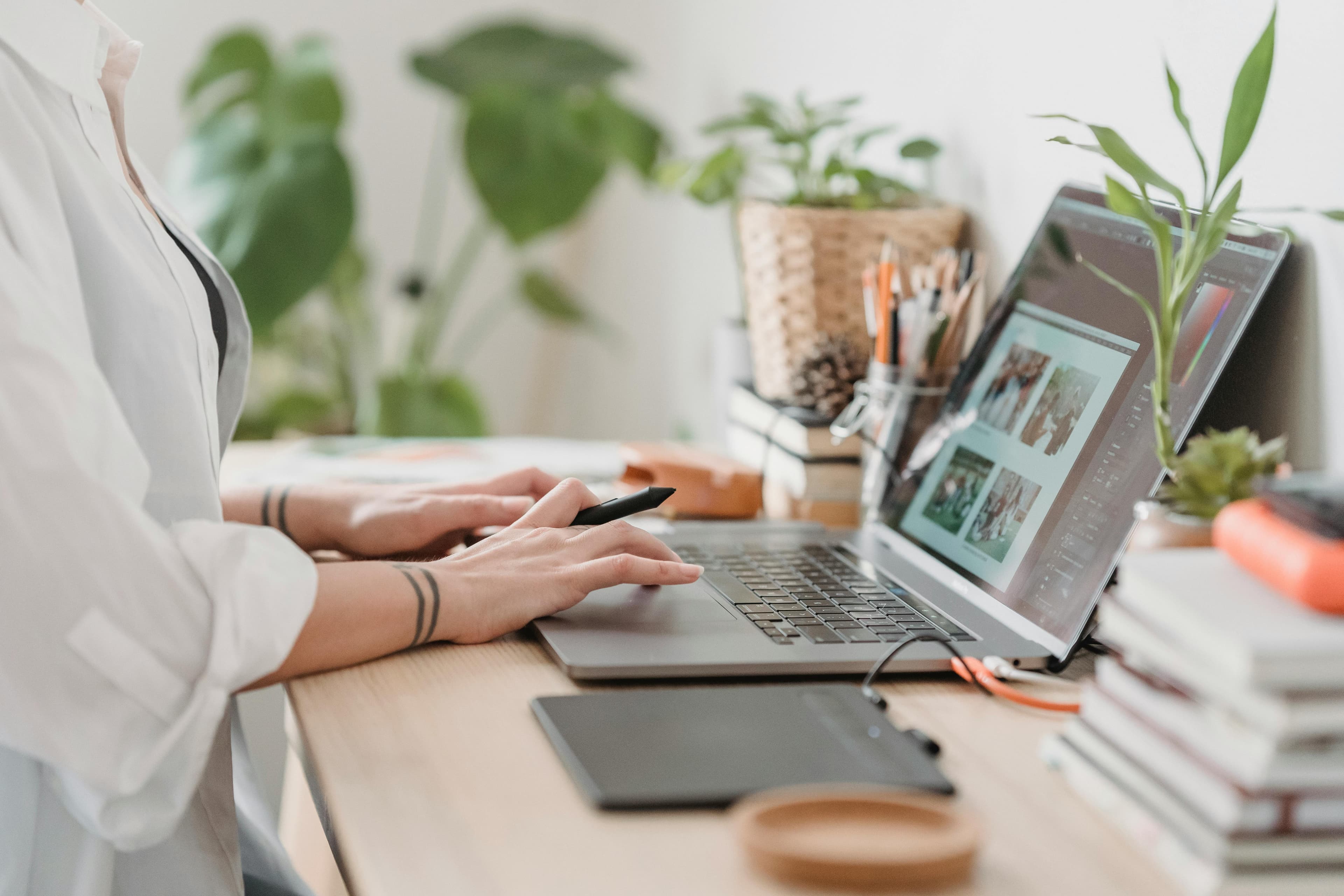 A person working at a desk with a laptop, holding a pen in hand while focusing on the screen displaying "Google PPC Ads". The workspace is adorned with potted plants, books, and various stationery items, creating a productive and organized environment.