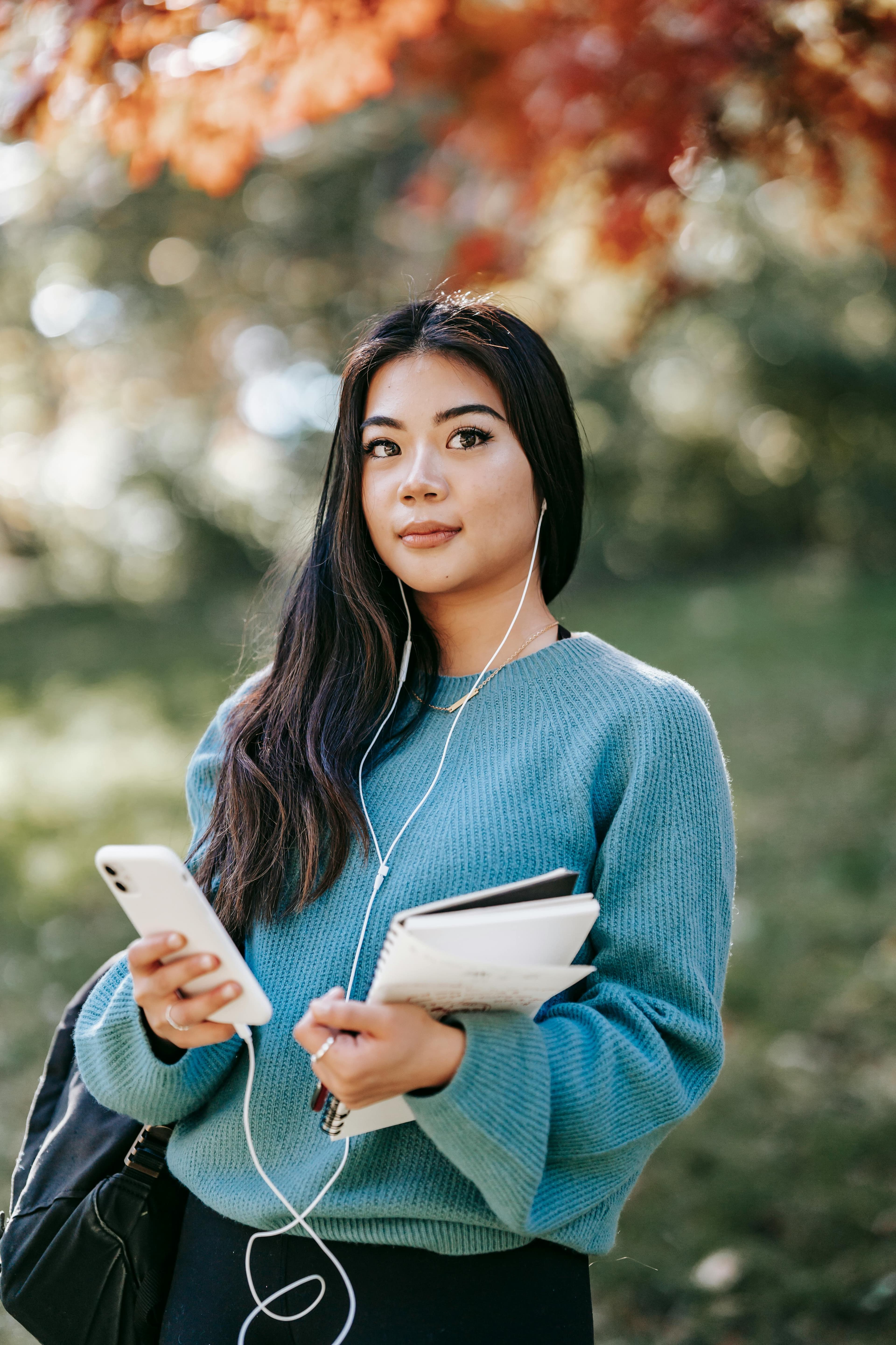 A young woman holding a smartphone and notebook, standing outdoors with autumn leaves in the background. This image conveys an approachable and thoughtful look, ideal for promoting "action pages" related to education or goal-setting.