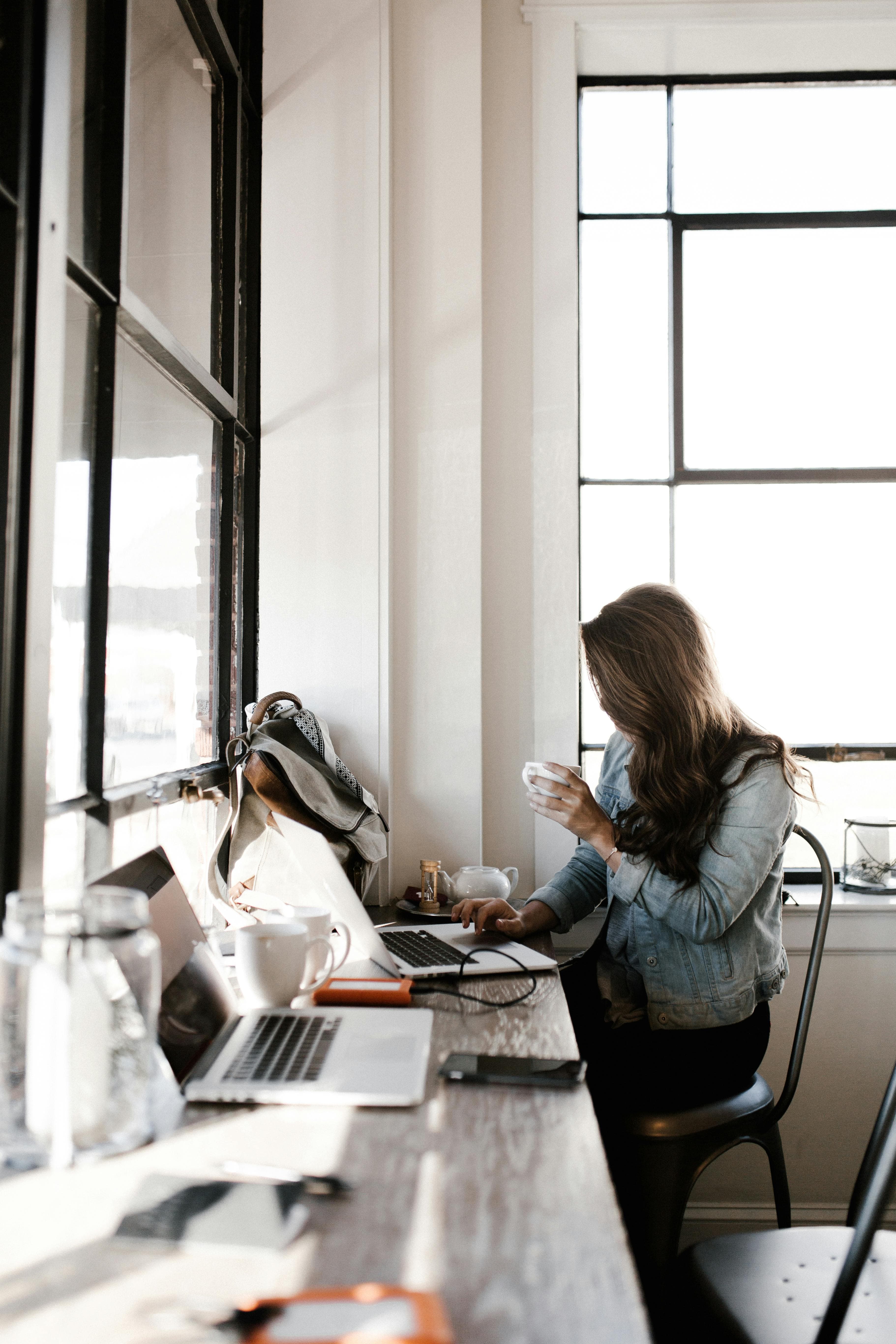 A woman works on her laptop at a bright cafe, holding a cup of coffee and surrounded by notebooks and devices. This image illustrates a professional setting, ideal for discussing the productivity benefits versus landing page cost investments.