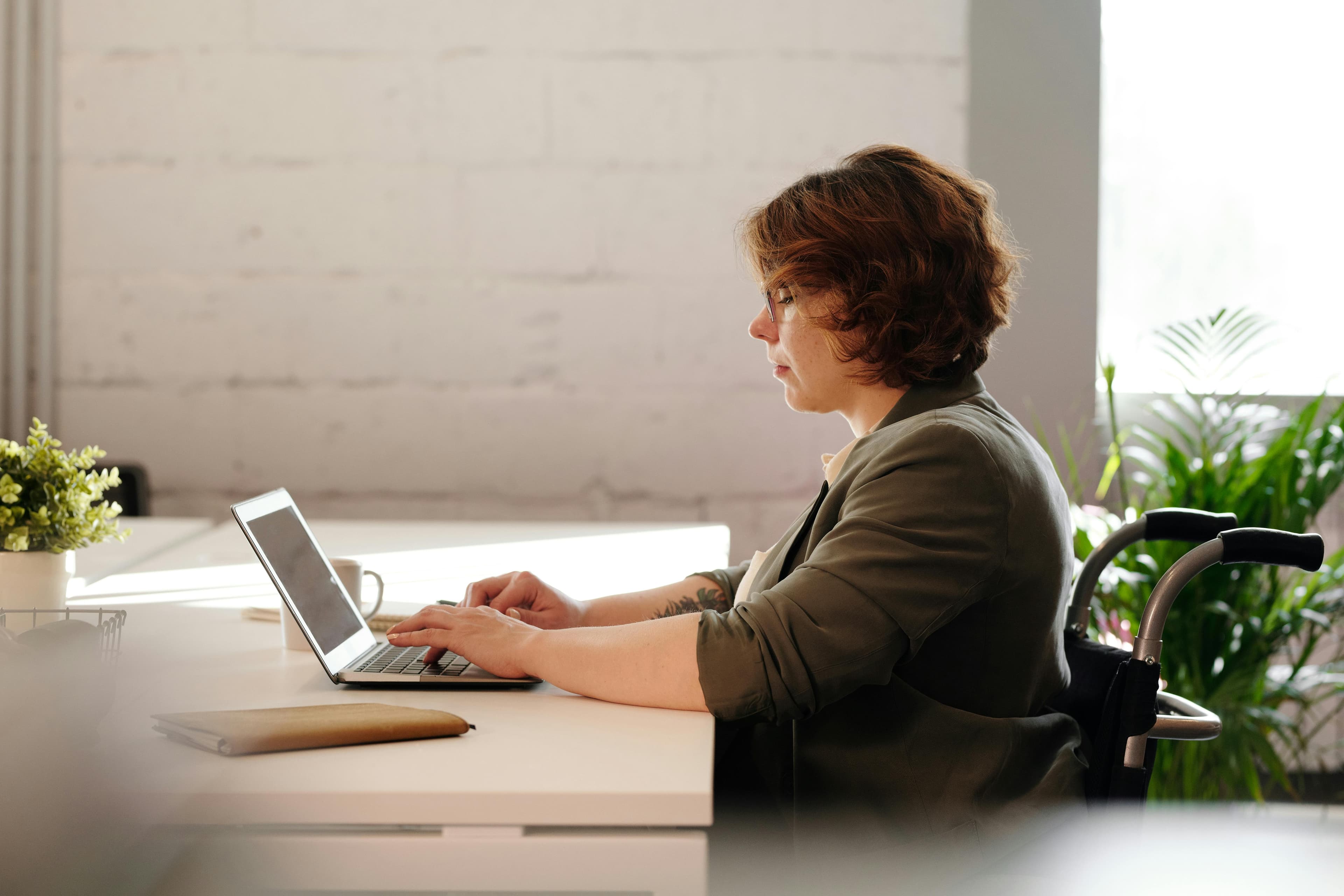 A woman with short brown hair and glasses sits at a desk, working on her laptop with focused concentration. She is in a bright office space with natural light streaming in and green plants in the background. The screen of her laptop displays a "Google PPC Ad" interface.