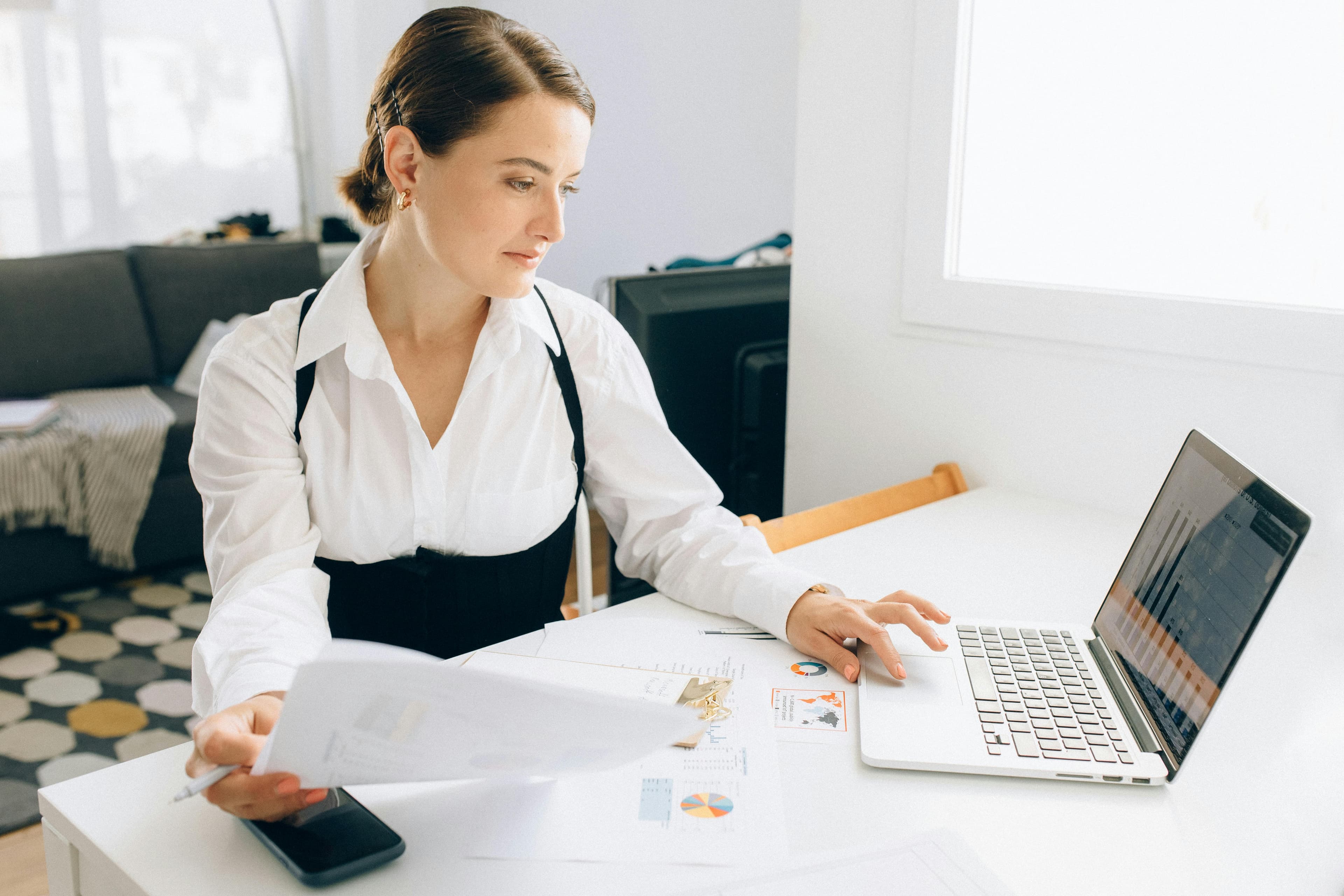 A professional woman at her desk, analyzing charts and documents while working on her laptop, representing the meticulous effort required to craft persuasive call to action phrases for targeted audiences.