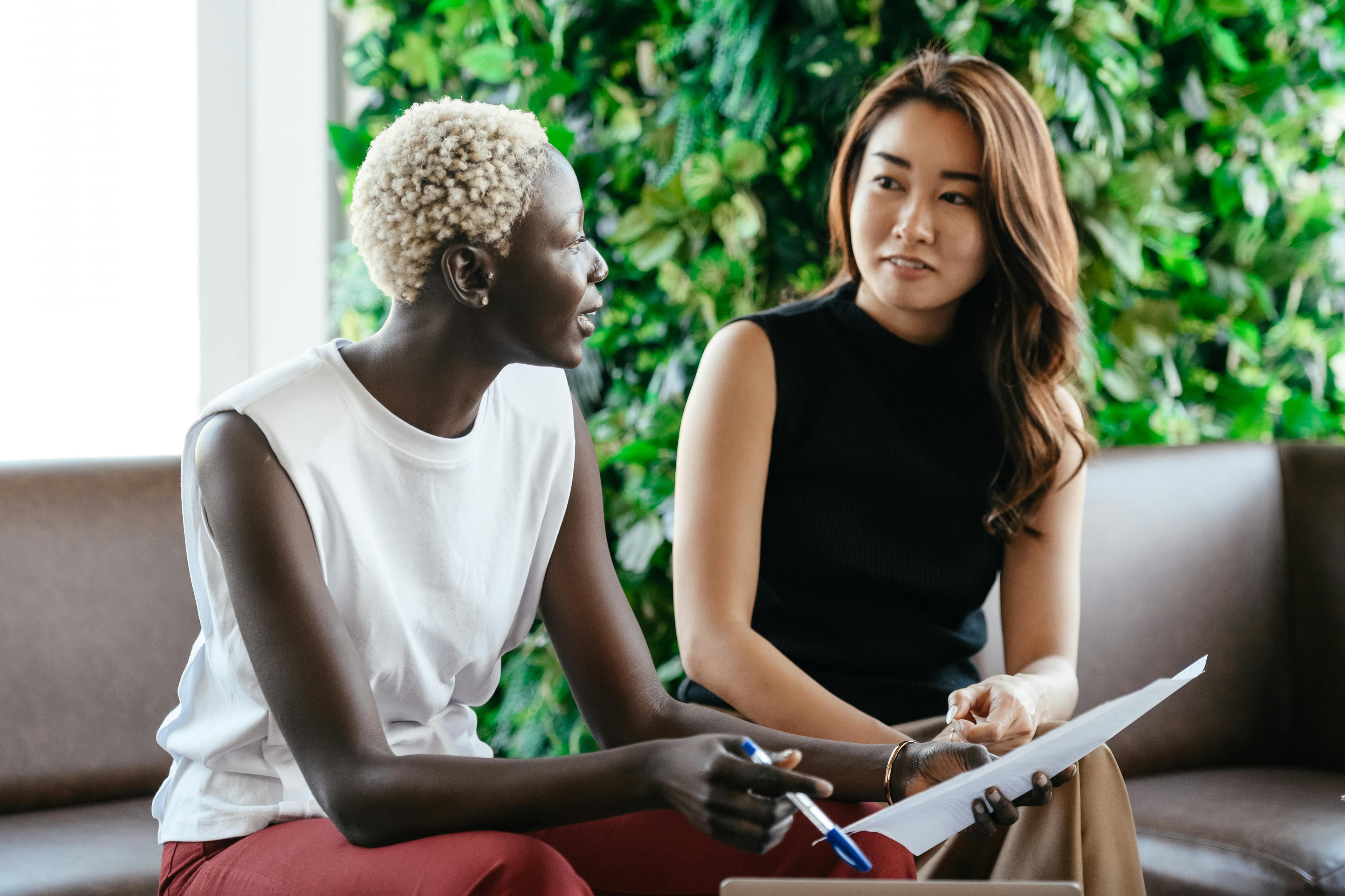 Two individuals sitting on a couch in a vibrant space with green foliage in the background, engaged in a discussion while reviewing a document. Perfect for "landing pages ideas" related to team collaboration, coaching, or creative brainstorming.