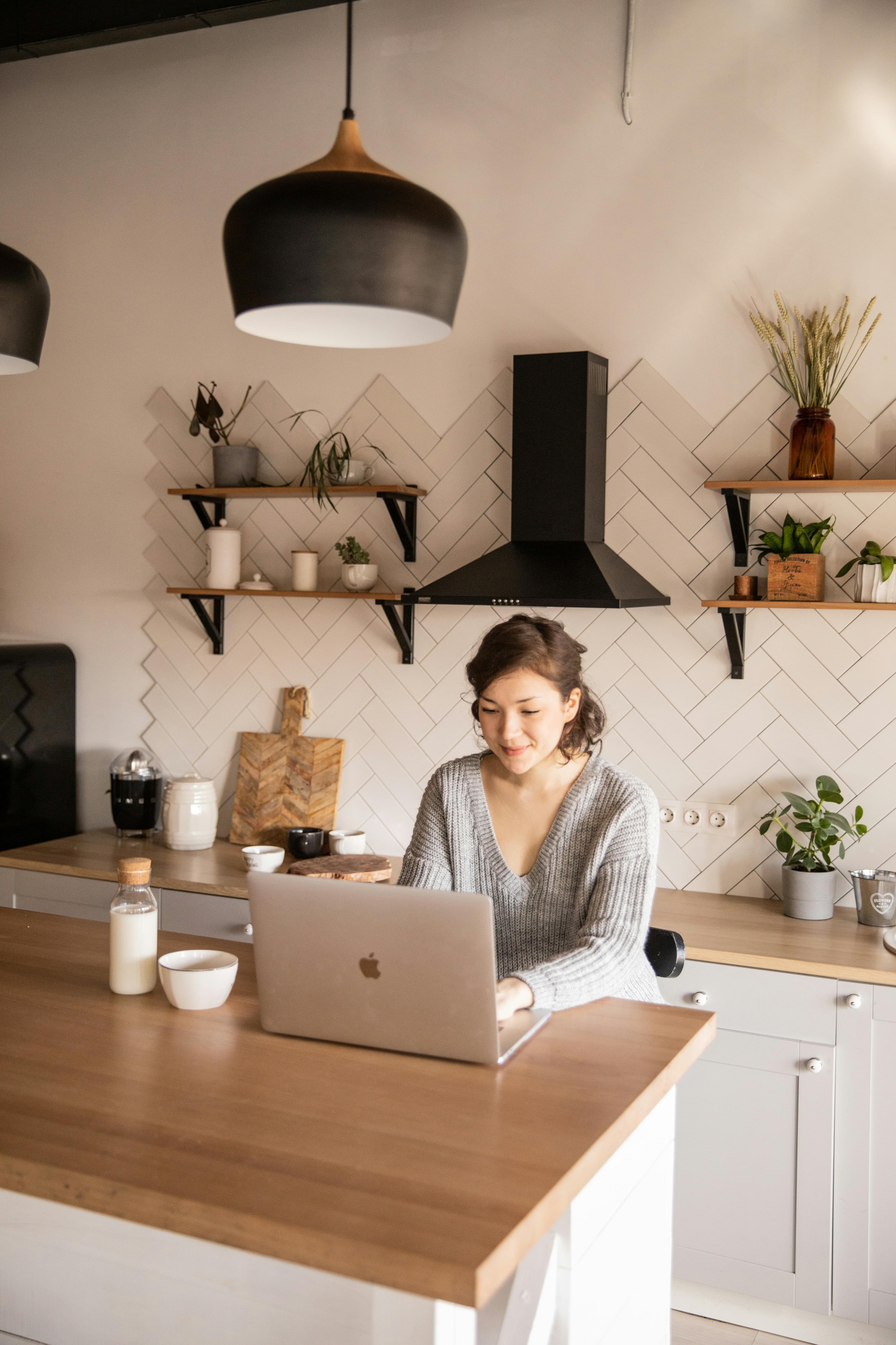 A serene kitchen scene featuring a smiling individual typing on a laptop at a wooden counter, surrounded by modern decor, highlighting the creative ambiance enhanced by an "AI persuasive writing tool."