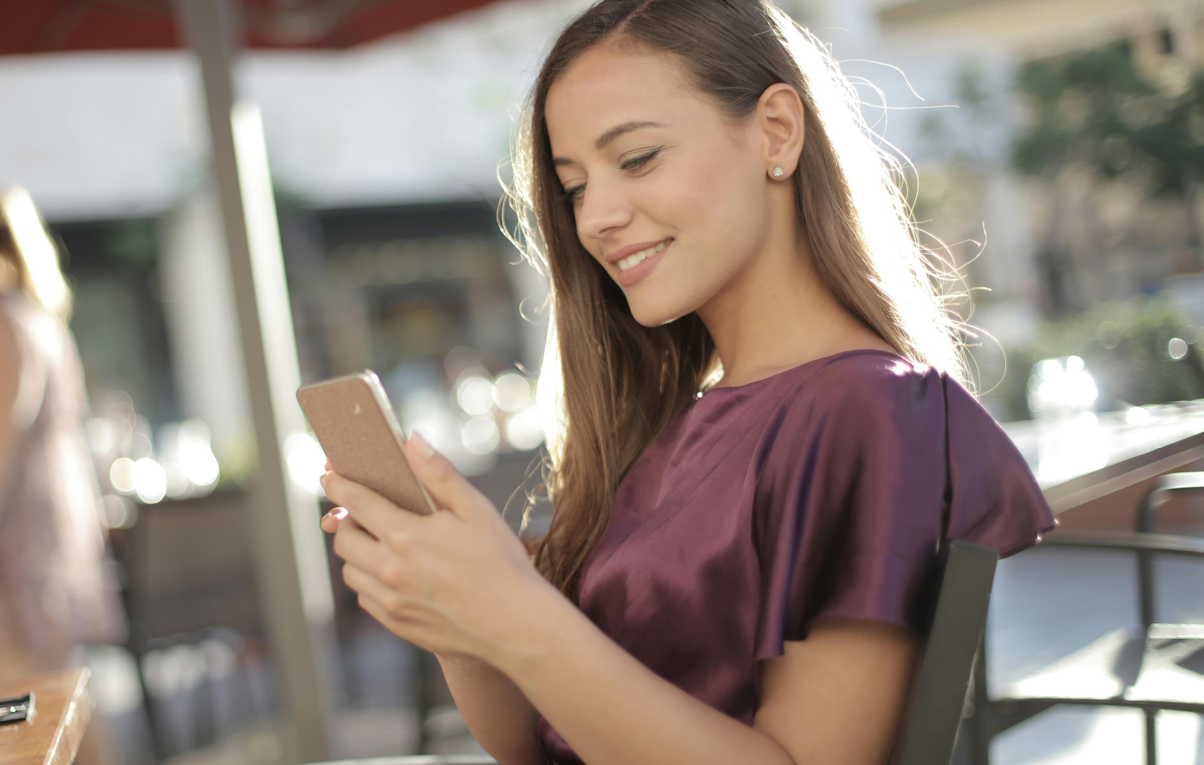 A young woman smiling while browsing product landing pages on her smartphone in a relaxed outdoor setting, enjoying a seamless mobile shopping experience.