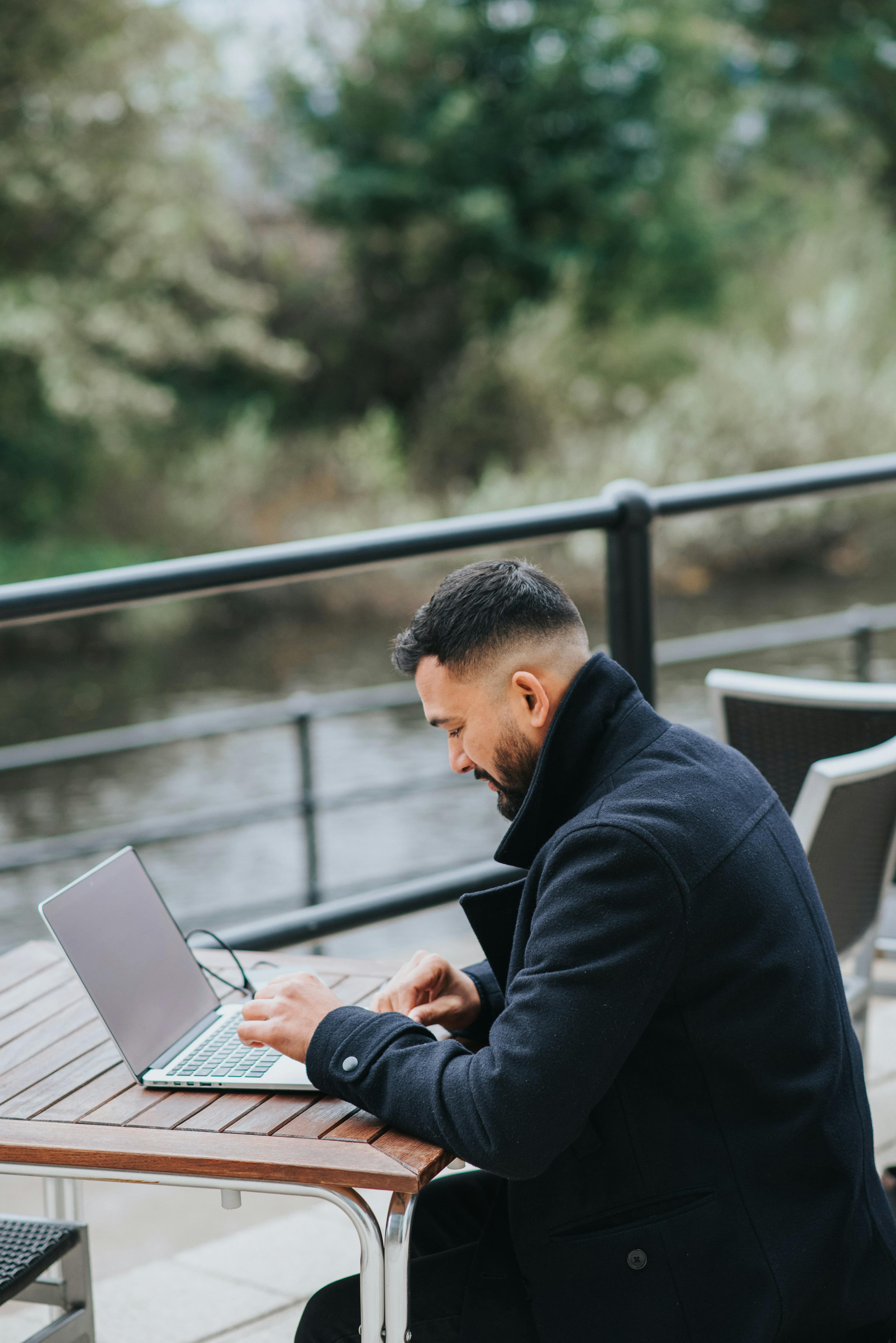 A man sits at an outdoor table, smiling as he types on his laptop. This relaxed and focused environment conveys how minimal landing page cost can enable efficient remote work setups.