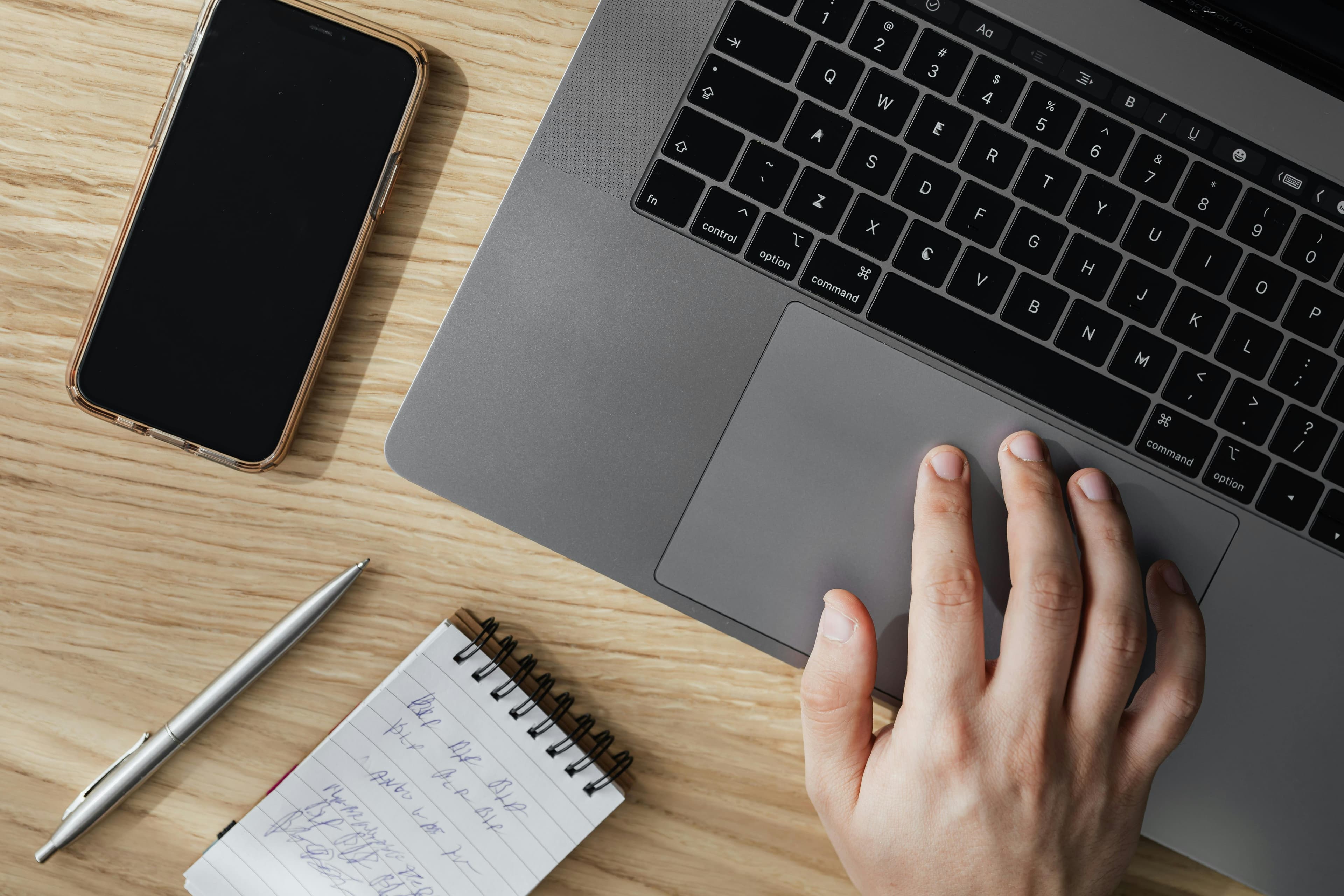 A close-up view of a person's hand on a laptop trackpad, with a smartphone and notebook nearby. This image highlights the attention to detail and precision often associated with management roles in the "leading vs management" debate.