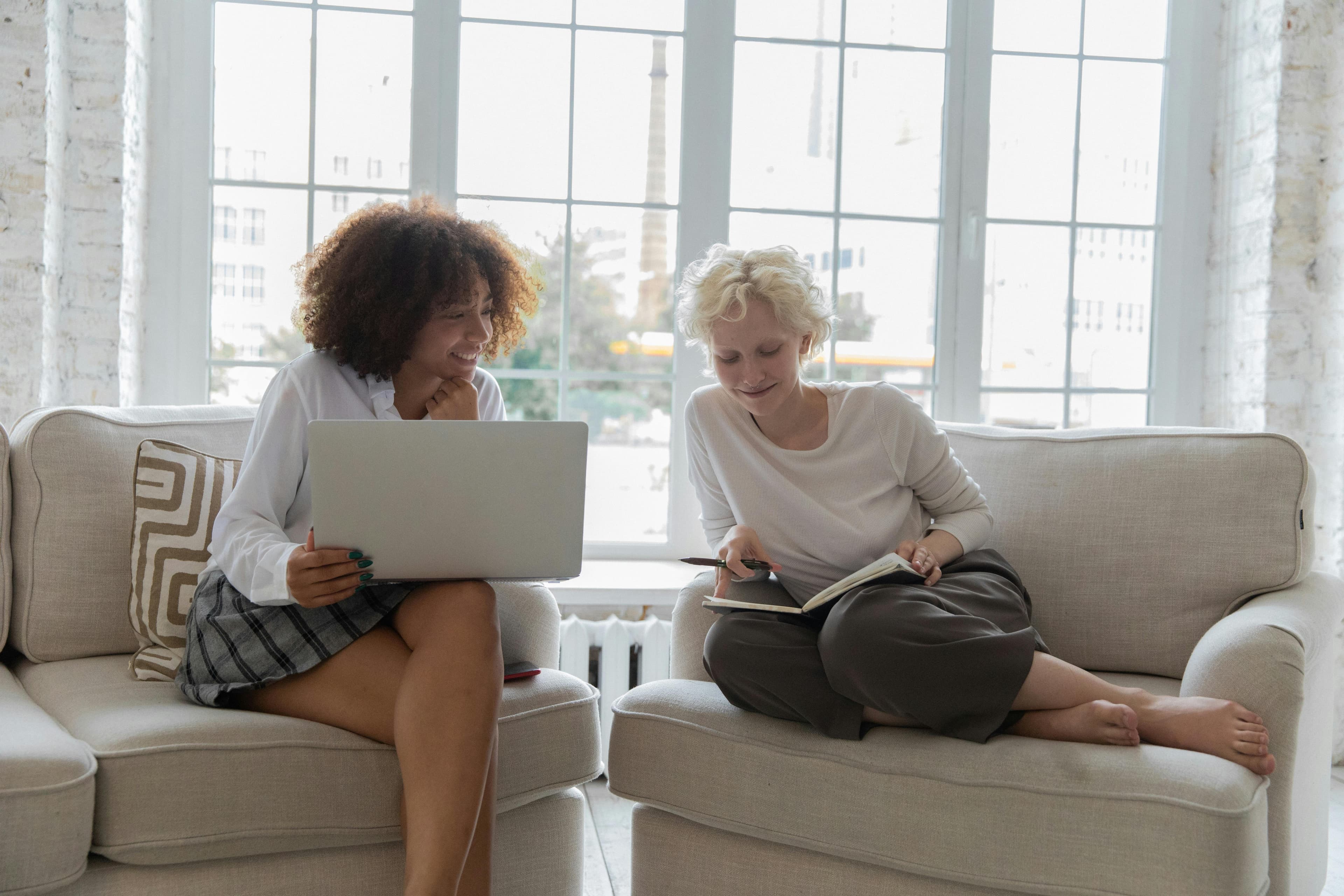 Two women sitting on comfortable sofas in a bright room, one working on a laptop and the other reading a notebook, discussing strategies for an inbound marketing funnel.