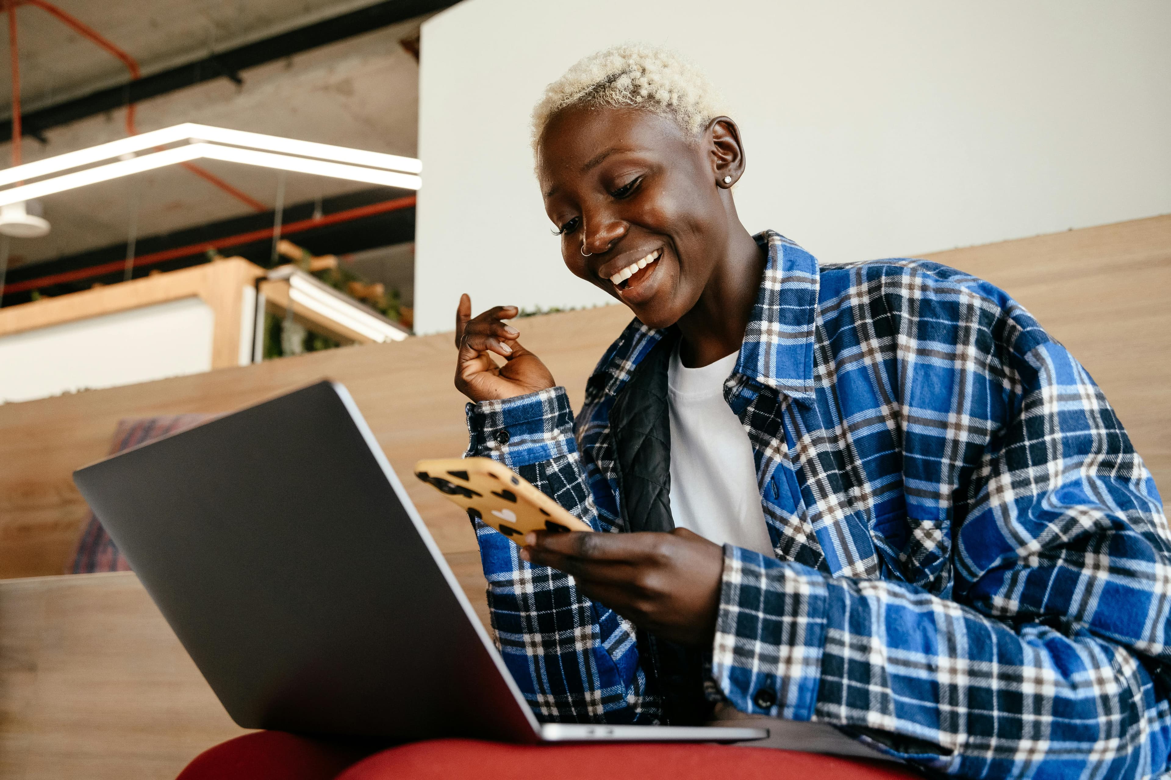 A person is sitting in a modern setting, smiling as they look at their phone while using a laptop. They are wearing a blue plaid shirt. This scene conveys a moment of joy and engagement, possibly related to exploring the app market on Chrome. The setting includes stylish, contemporary lighting and a cozy, casual atmosphere.