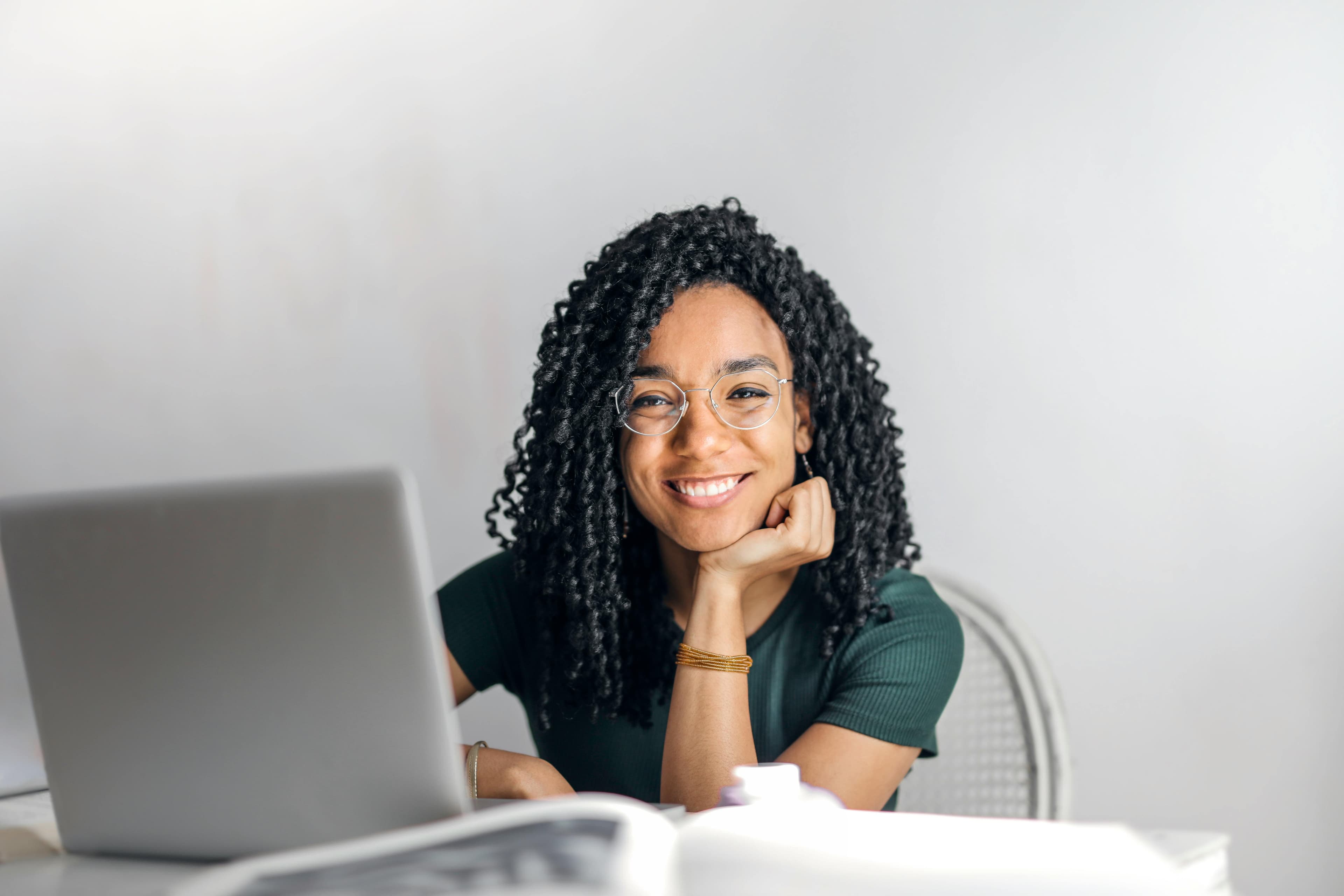 A woman with curly hair and glasses, smiling while working on her laptop in a bright, minimalist space. She appears engaged and focused, possibly working on a page design project.