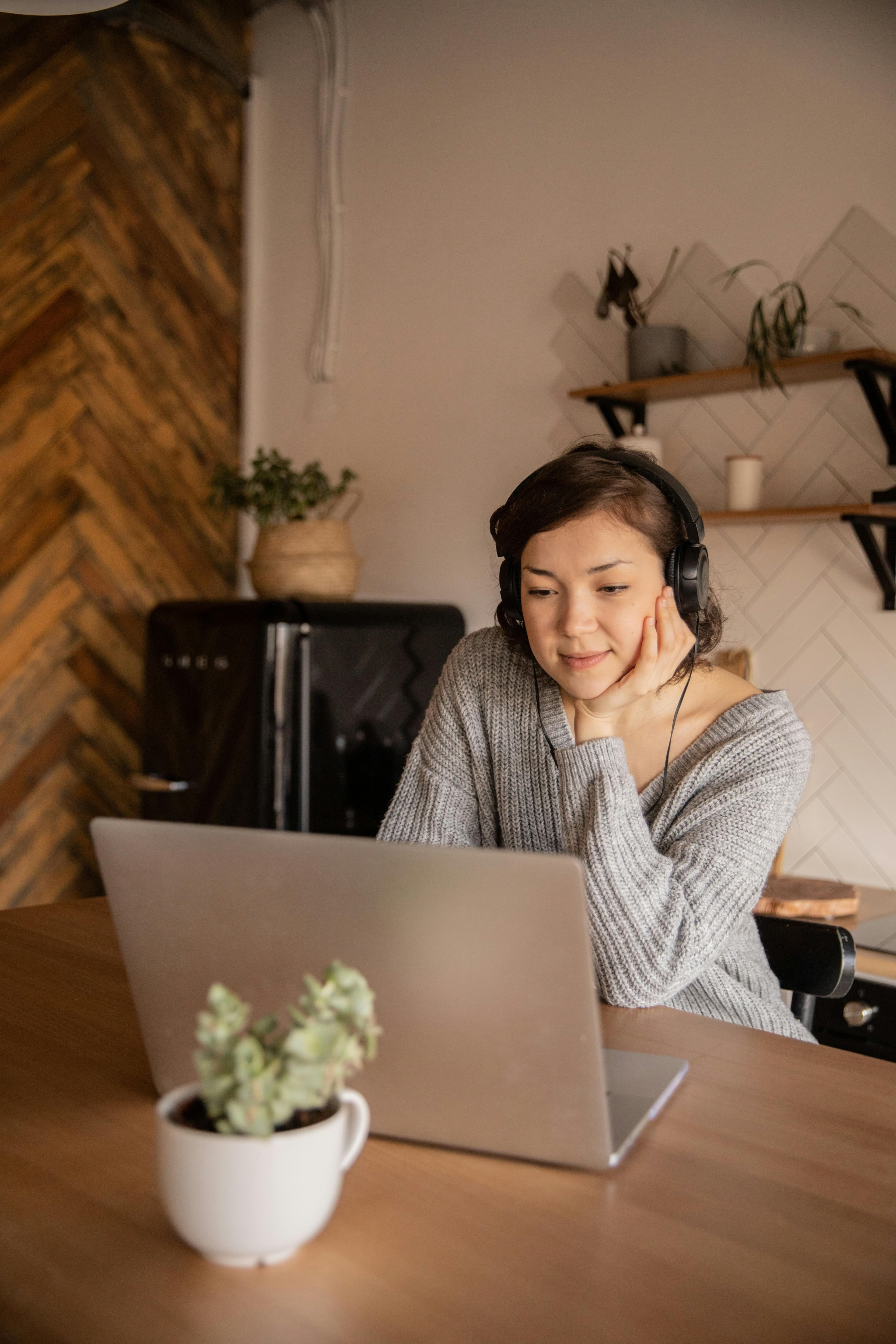 A woman wearing headphones leans on her hand as she attentively watches her laptop screen in a cozy kitchen environment, showcasing focus and creativity in "automated content creation."