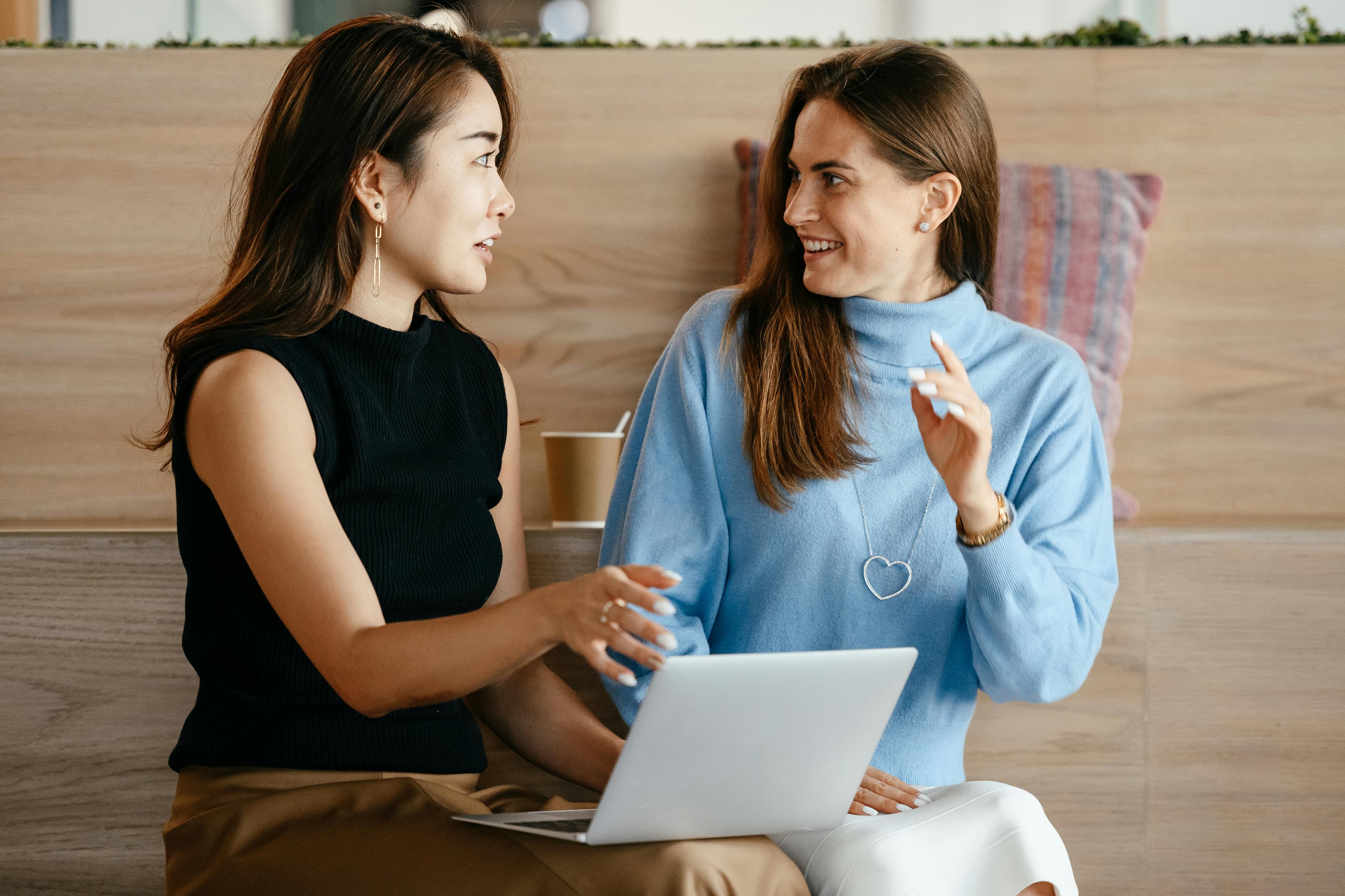  Two women sitting together with a laptop, engaged in a lively discussion in a modern, cozy environment. Great for "landing pages ideas" centered on teamwork, mentorship, or casual workspaces.