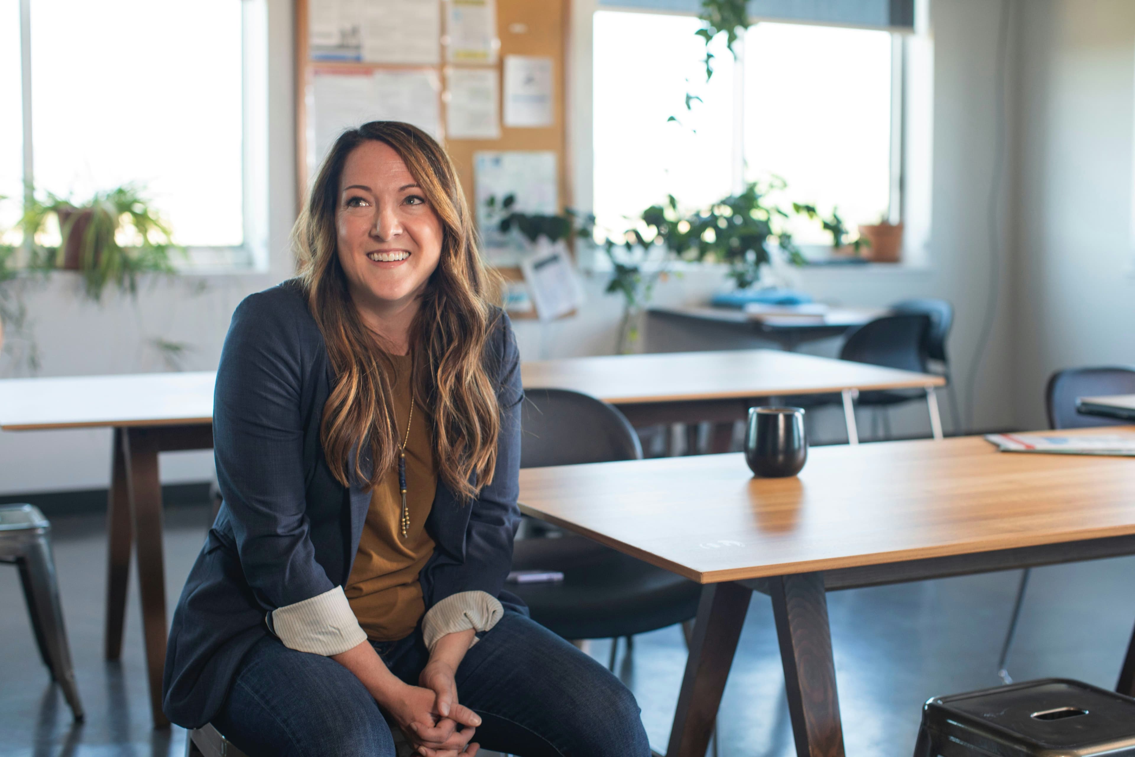 A woman with long brown hair and a friendly smile sits at a wooden table in a well-lit office with large windows and potted plants. The background features other tables, chairs, and a bulletin board. This setting is ideal for discussing and developing WooCommerce landing pages.