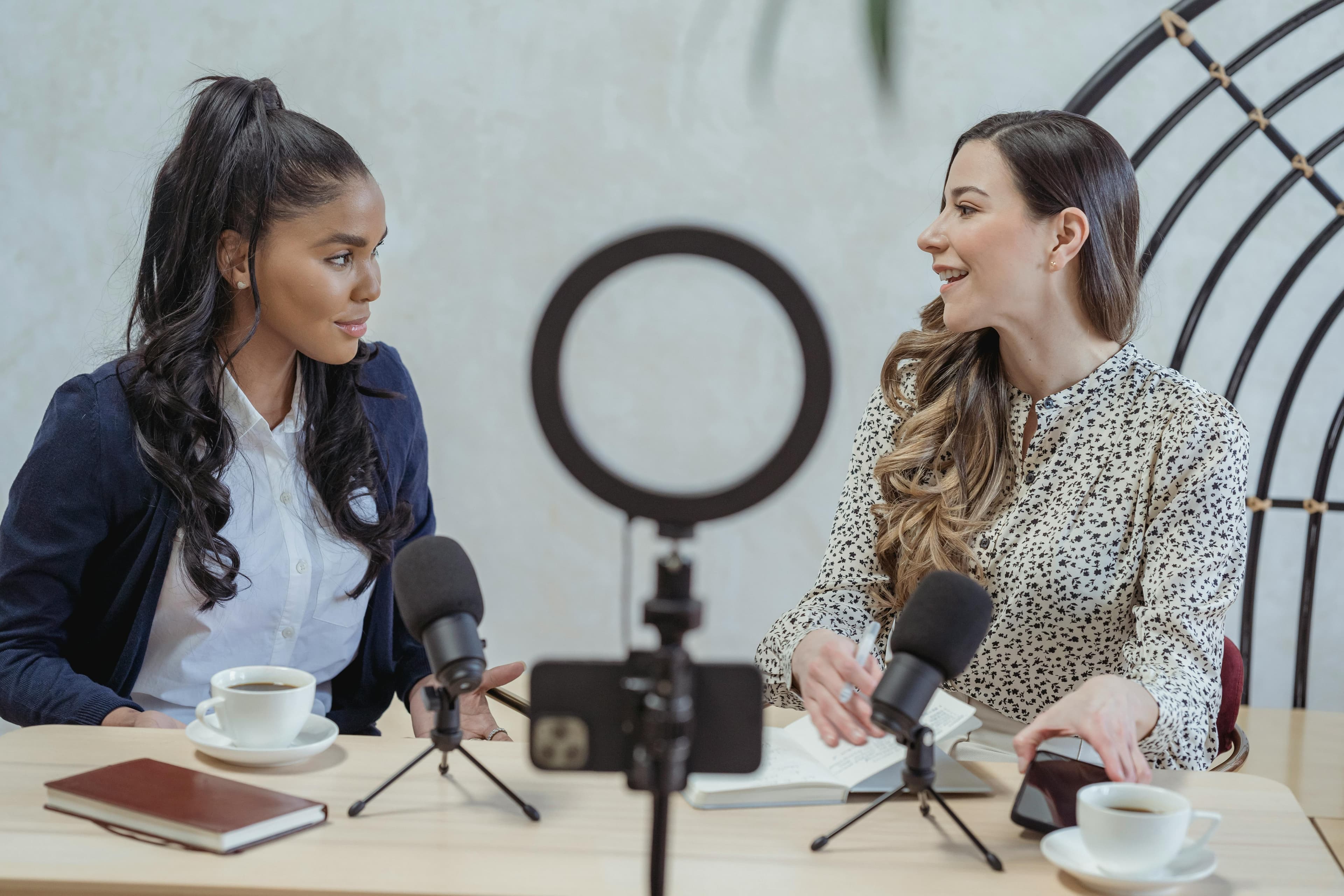 Two women sitting at a table with microphones and a ring light, engaging in a discussion, representing the work of influencer marketing agencies.