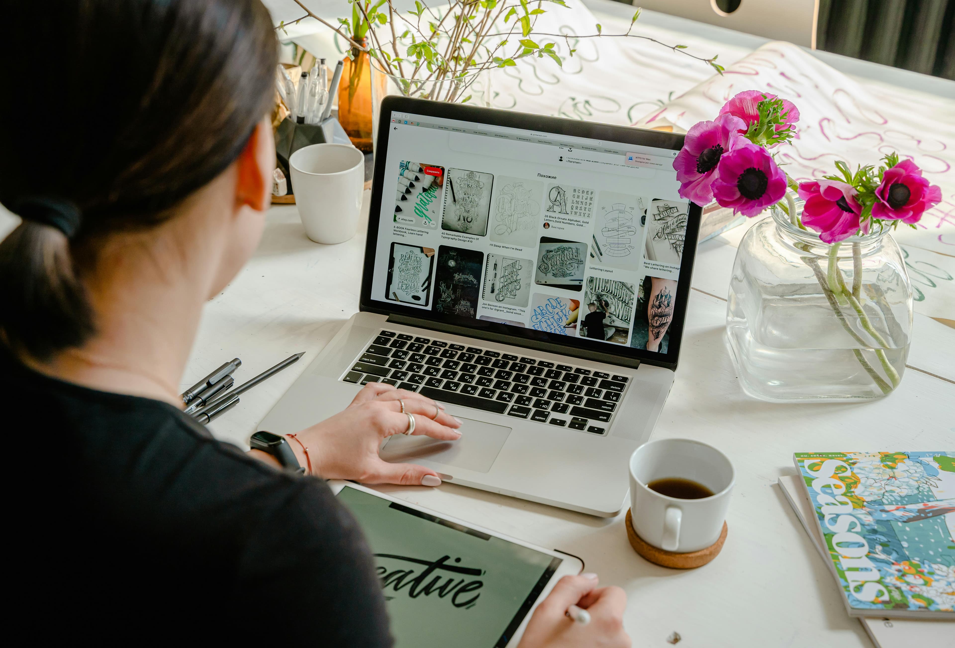 A person sitting at a desk browsing design inspirations on a laptop, with creative sketches displayed on the screen. The workspace is adorned with fresh flowers and coffee, showcasing the thoughtful and artistic process involved in crafting the best product landing pages for engaging and visually appealing user experiences