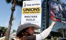 A woman stands on a sunny street holding a picket sign, while a Netflix billboard is visible in the background.