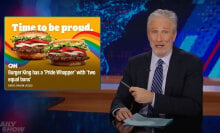 A man sits behind a talk show desk. In the top-left is a Burger King advertisement for Pride.